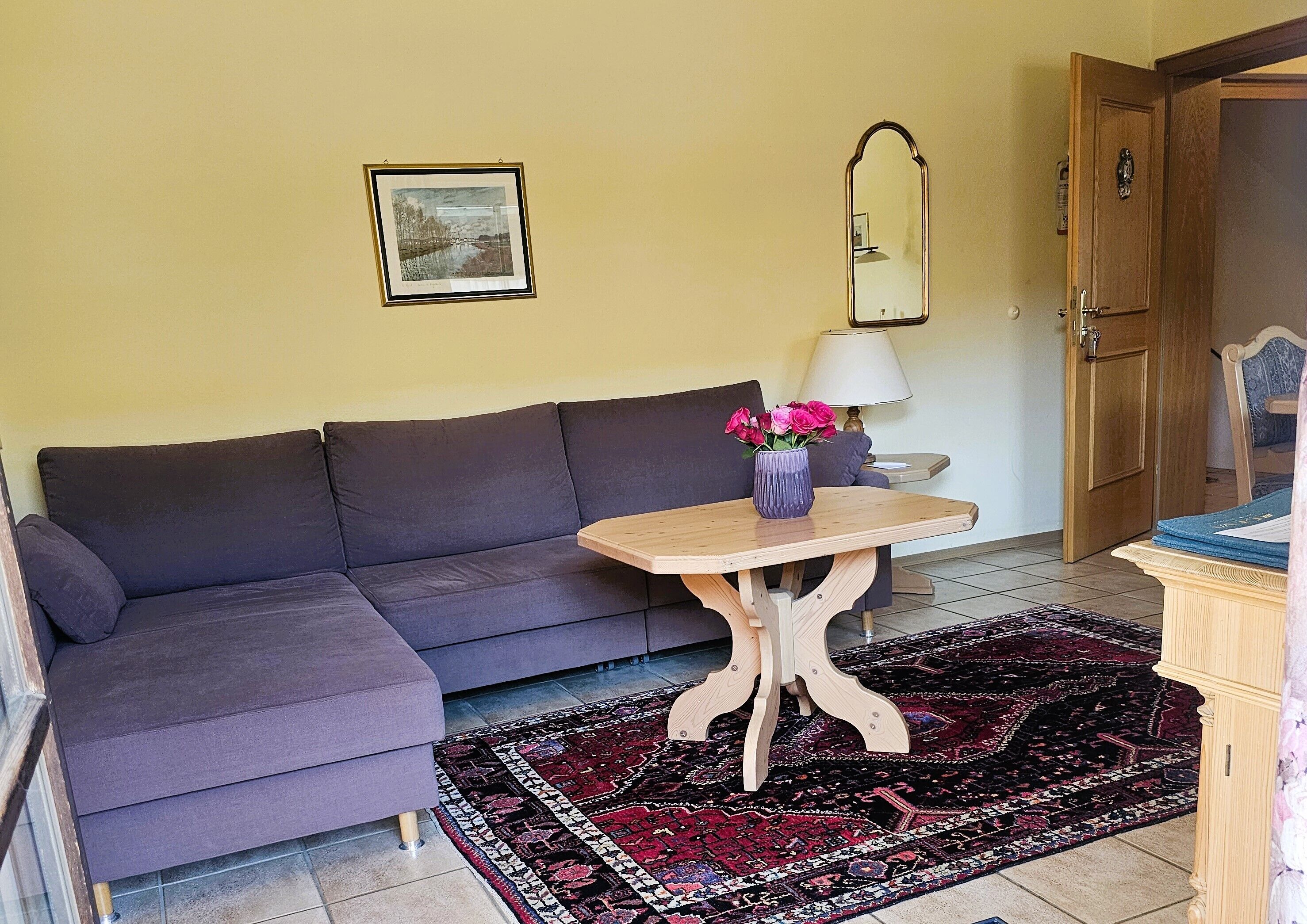 A room with a dark-colored sofa, a small wooden table with a vase of flowers, a patterned rug, a lamp, and a mirror on the wall.