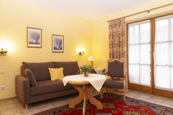 A living room with a sofa, a table with a vase of flowers, a lamp, and a chair. There are two framed pictures on the wall and a window with curtains.