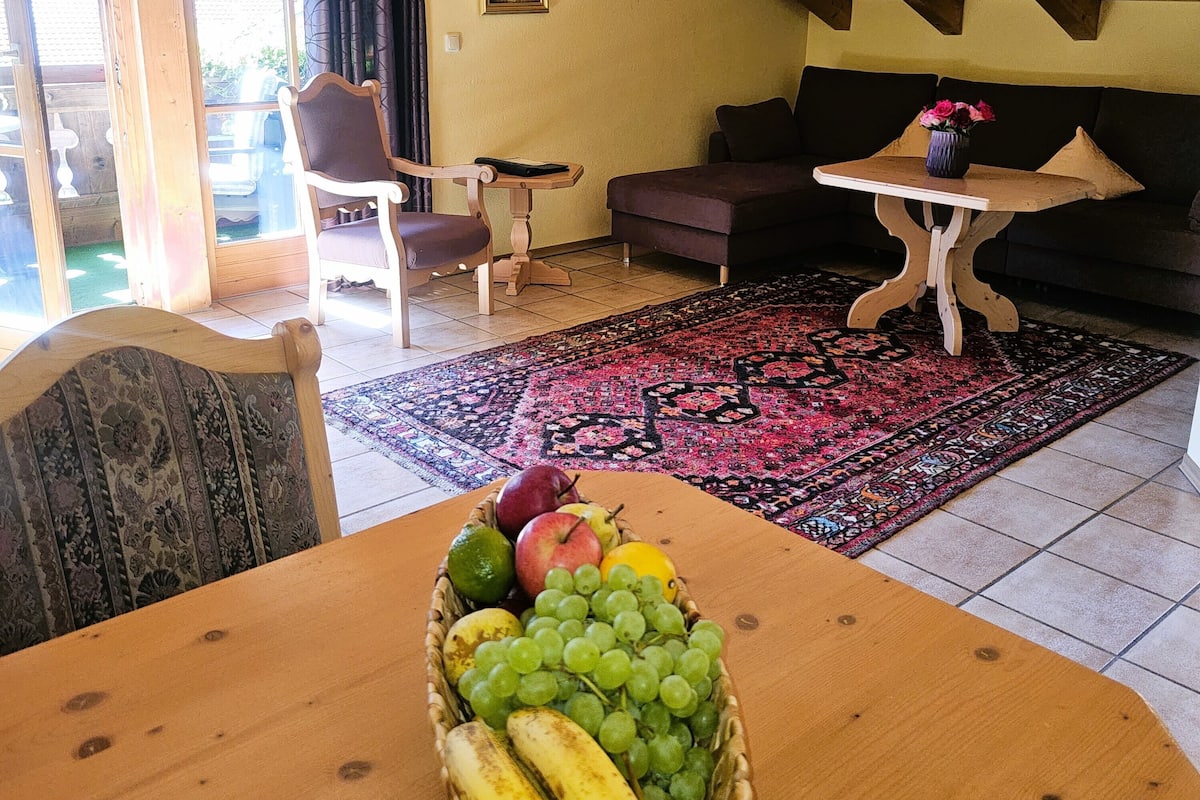 Ein gemütliches Zimmer mit Holzbalkendecke, einem Esstisch mit Obstkorb, einer Couch und einem kleinen Tisch mit einer Vase.