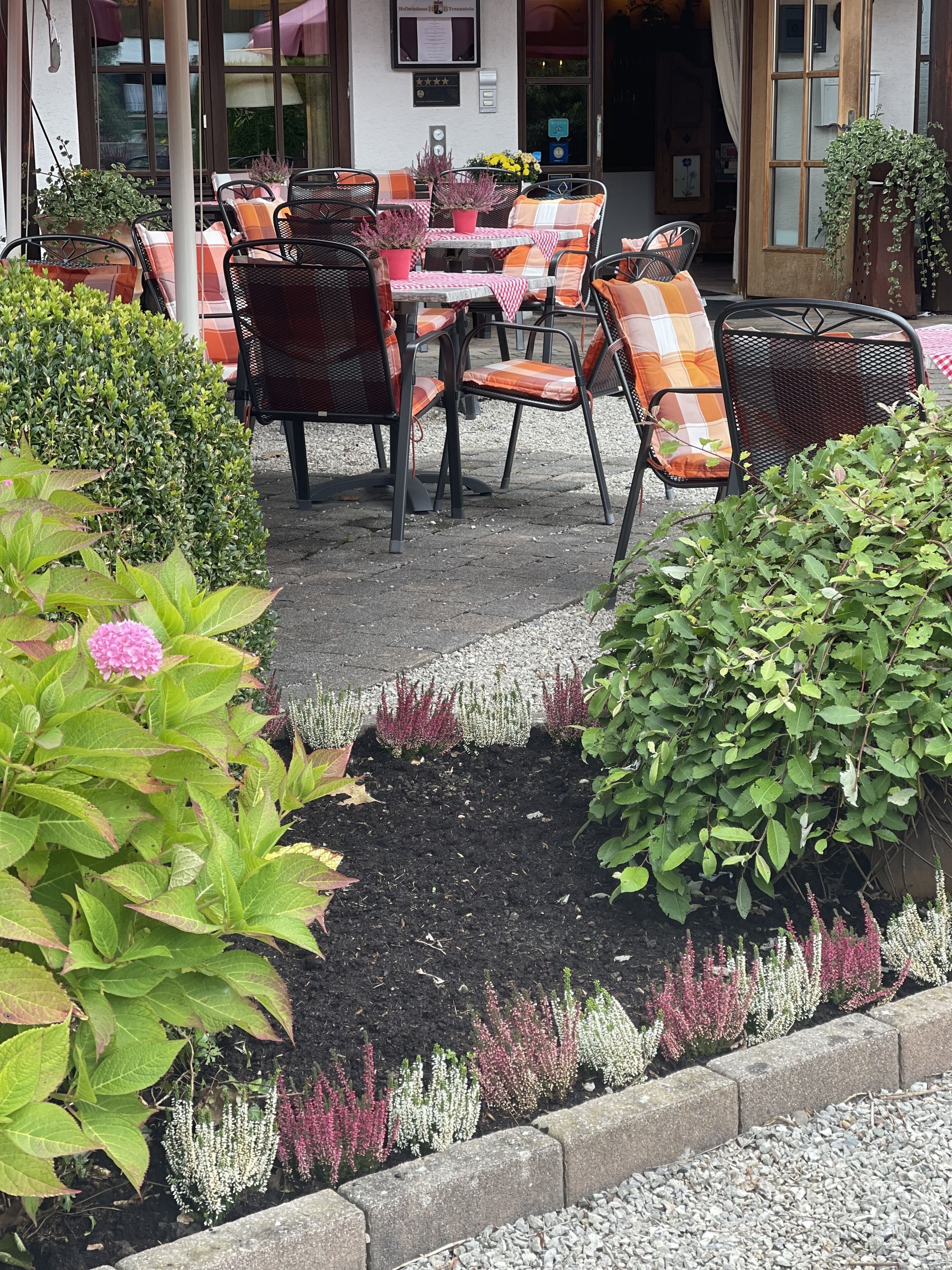 An outdoor seating area with tables and chairs, surrounded by potted plants and a flower bed.