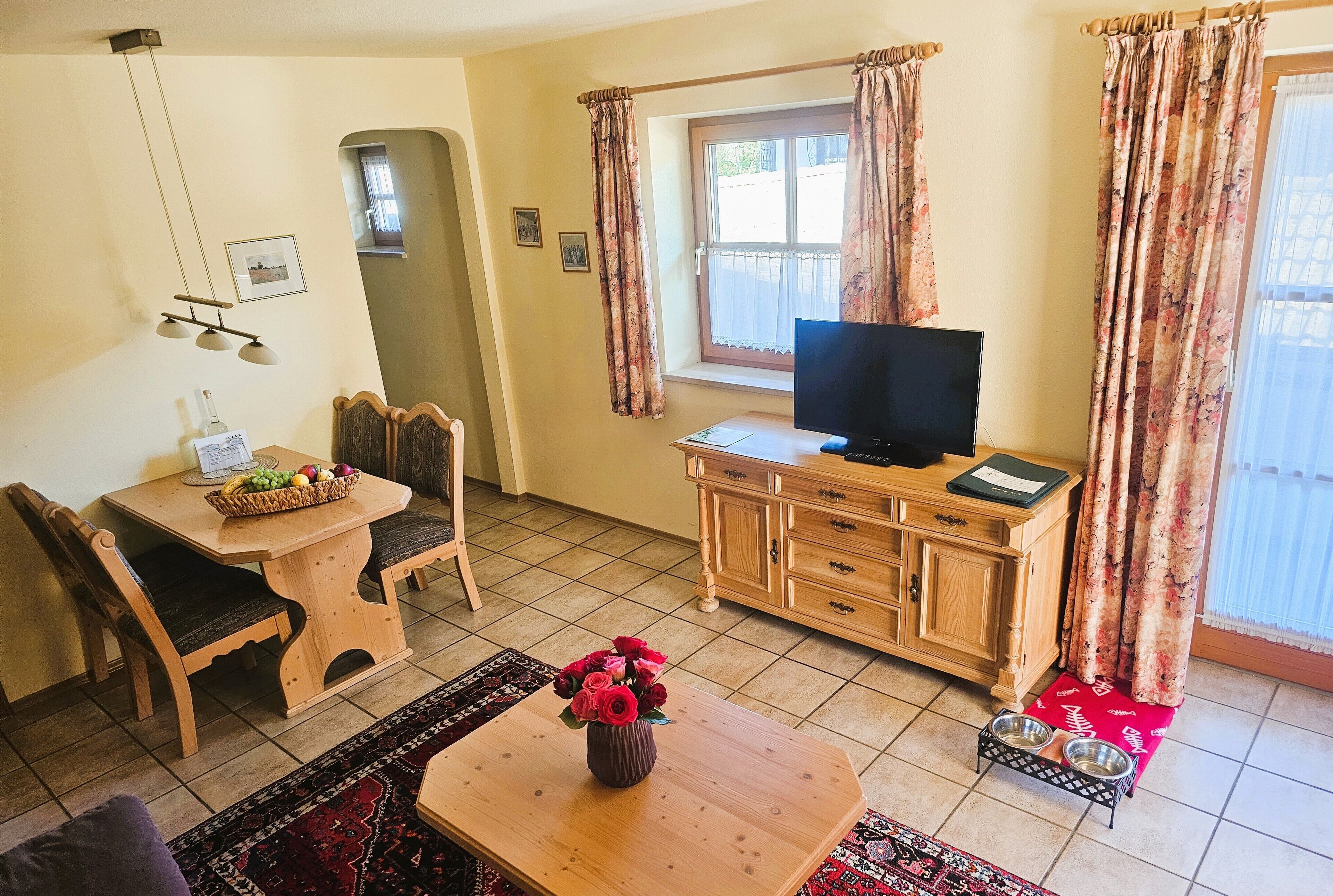 A living room with a wooden dining table, chairs, a TV on a wooden cabinet, and a window with curtains.