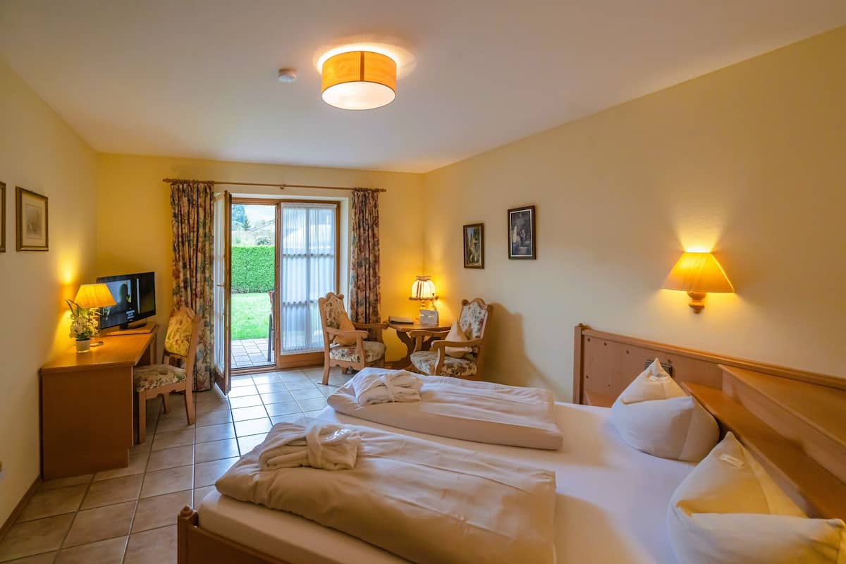 Ein Hotelzimmer mit zwei Betten, einem Schreibtisch mit Fernseher, einem Sessel, einem Tisch mit Lampe und Blick ins Freie.