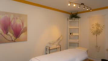 A room with a bed, a massage table, shelves with plants, and artwork on the walls.