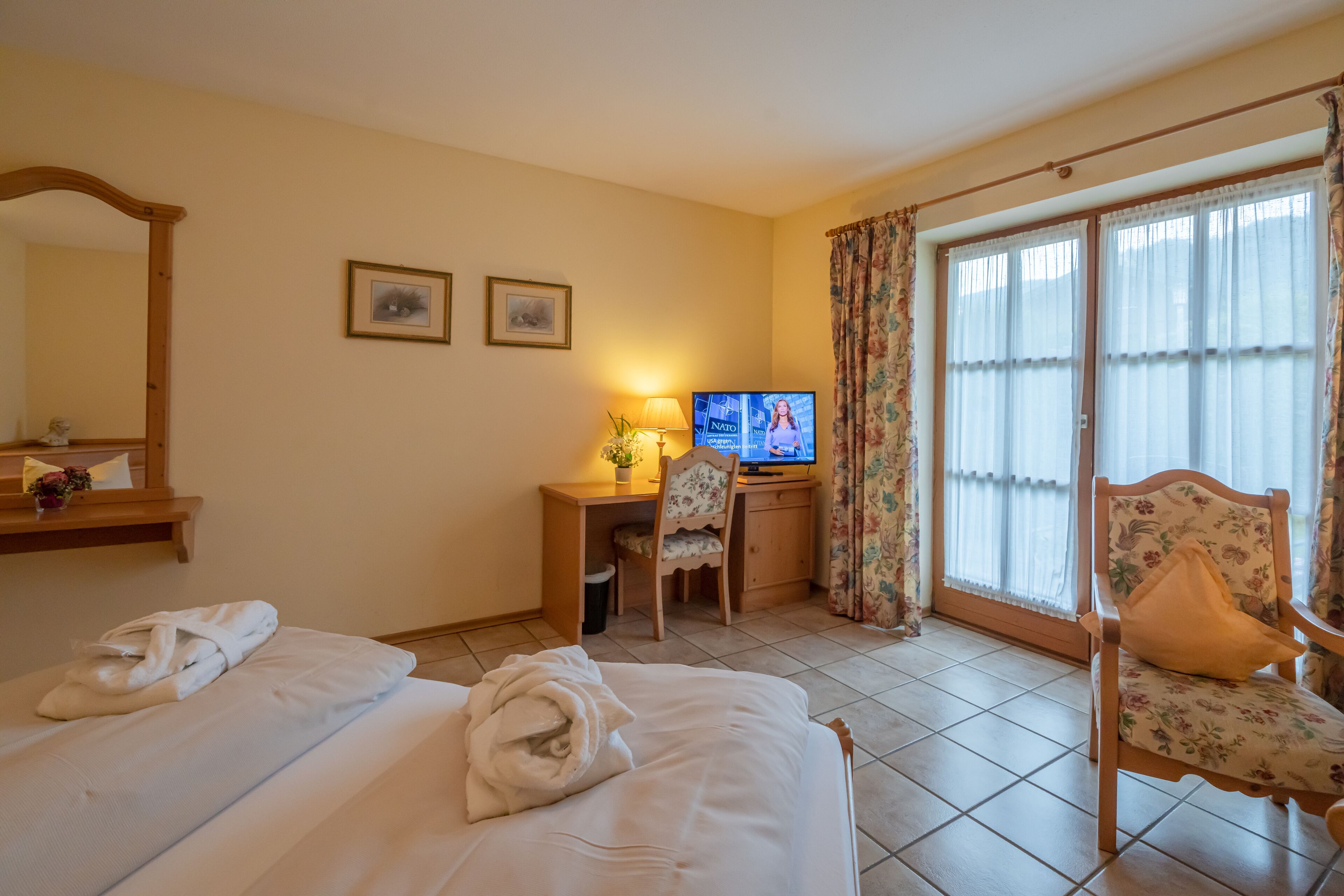 A hotel room with a bed, a desk with a television, a chair, and a window with curtains.
