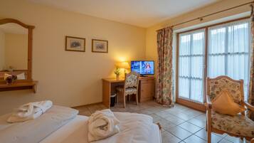 Quarto de hotel com cama, escrivaninha com televisão, uma cadeira e janela com cortinas.