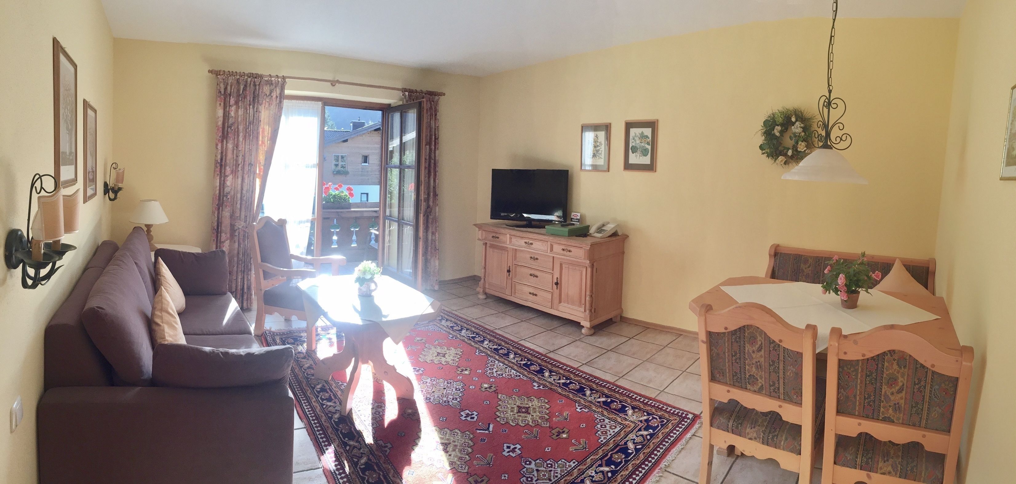 Ein Wohnzimmer mit Sofa, Fernseher, Esstisch mit Stühlen, einem gemusterten Teppich und einem Balkon mit Vorhängen.