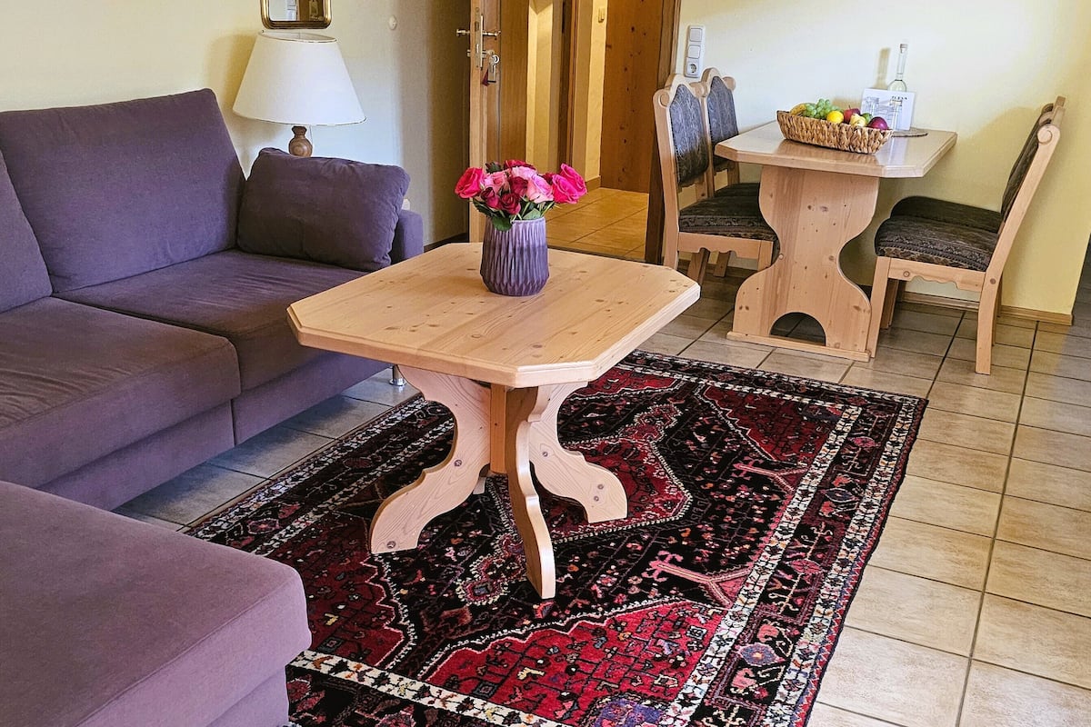 Ein Wohnzimmer mit einem violetten Sofa, einem Holztisch mit Blumenvase, einem gemusterten Teppich und einem Holzesstisch mit Stühlen.