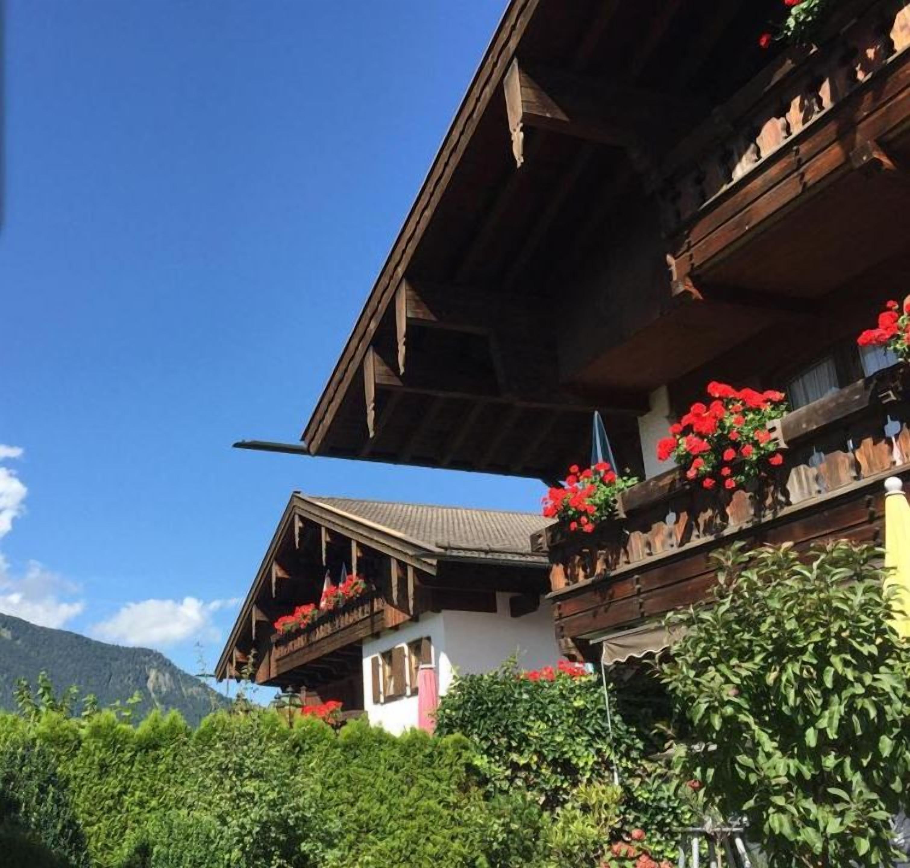 Un bâtiment de style chalet traditionnel, avec une façade en bois, des balcons ornés de fleurs rouges et un paysage montagneux en arrière-plan.