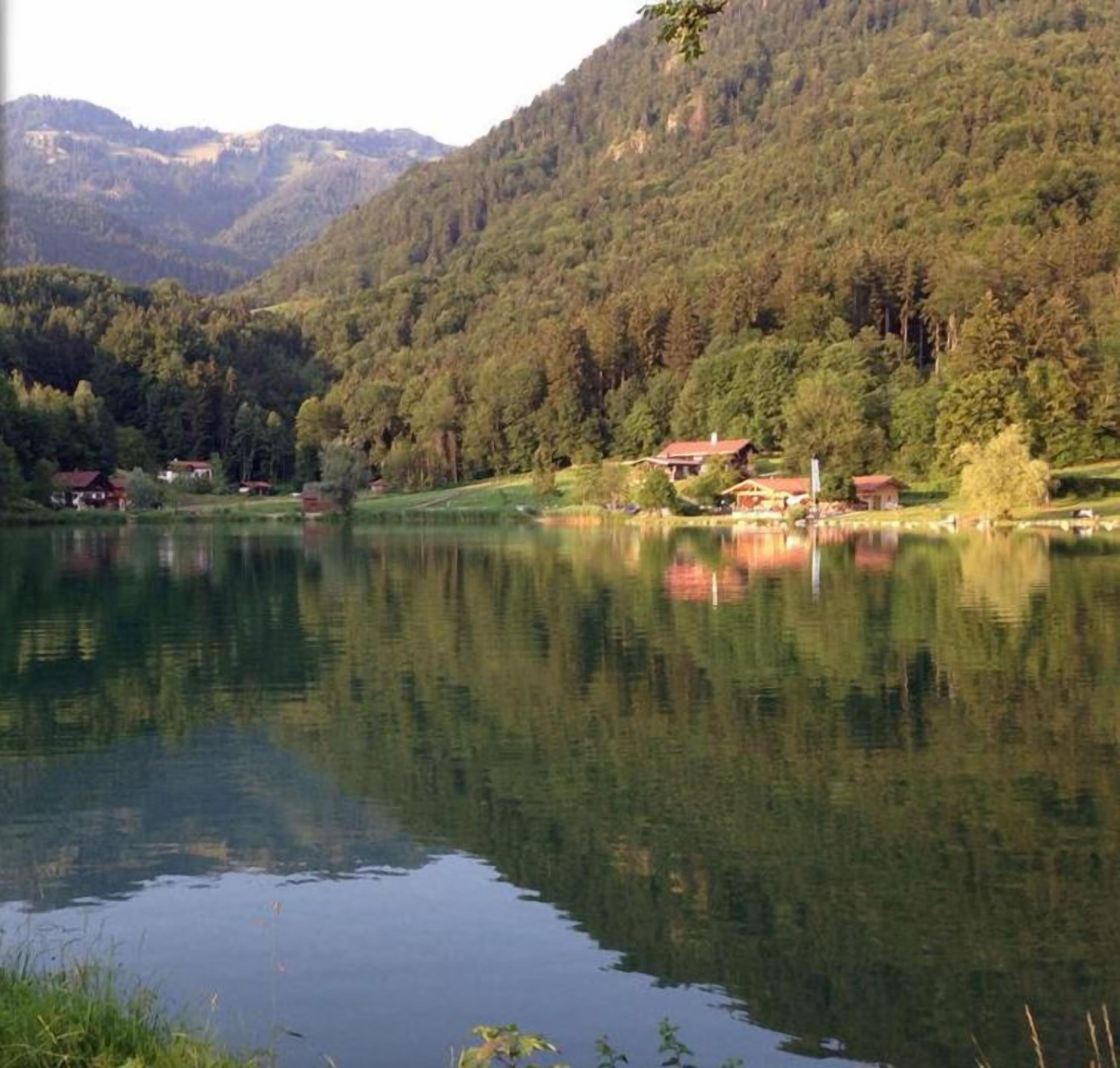 Un lago tranquillo circondato da colline boscose, con il logo dell'hotel 'Achentaler Vital Hotel' nell'angolo in alto a sinistra.