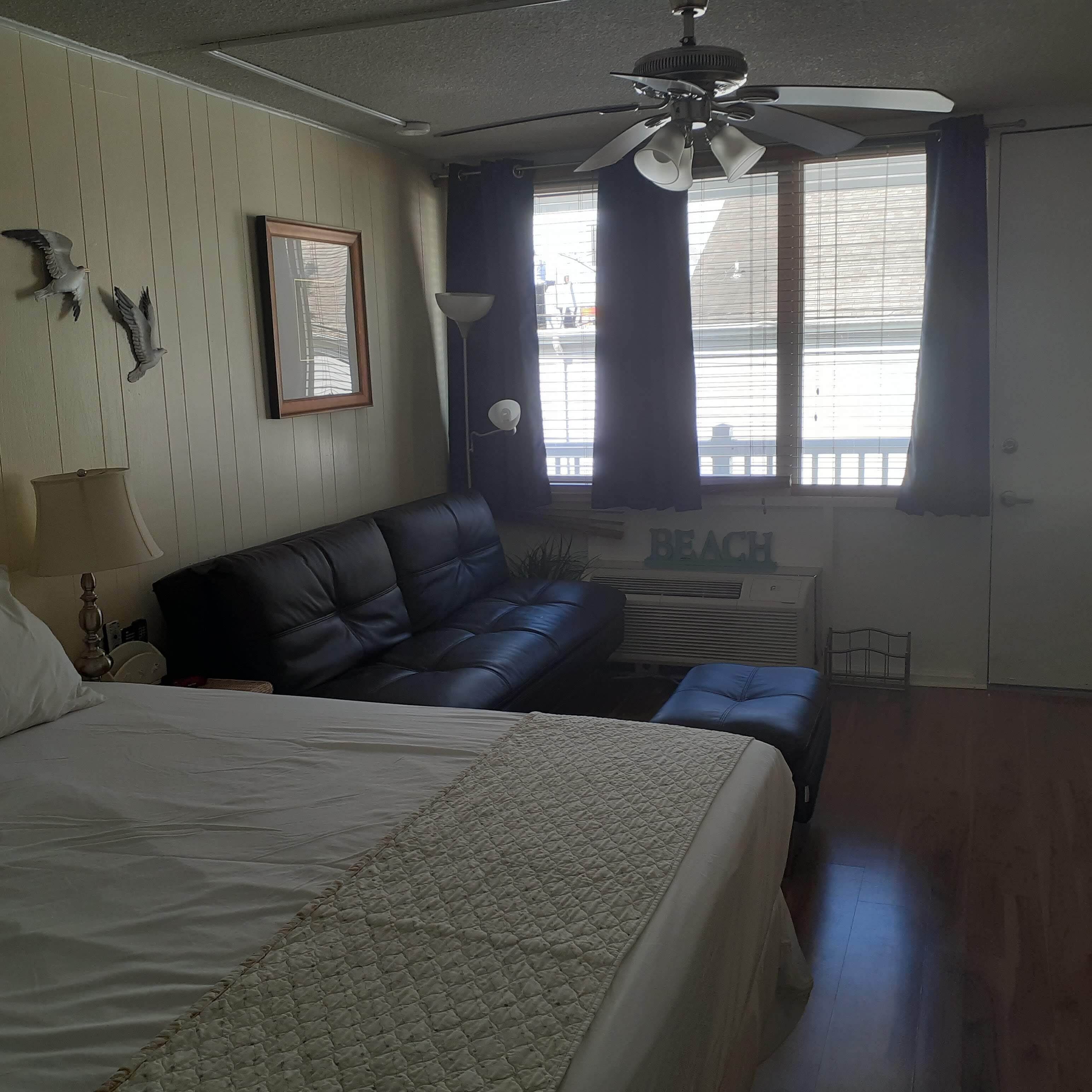 A bedroom with a bed, a sofa, a ceiling fan, a window with curtains, and a wall-mounted air conditioner.