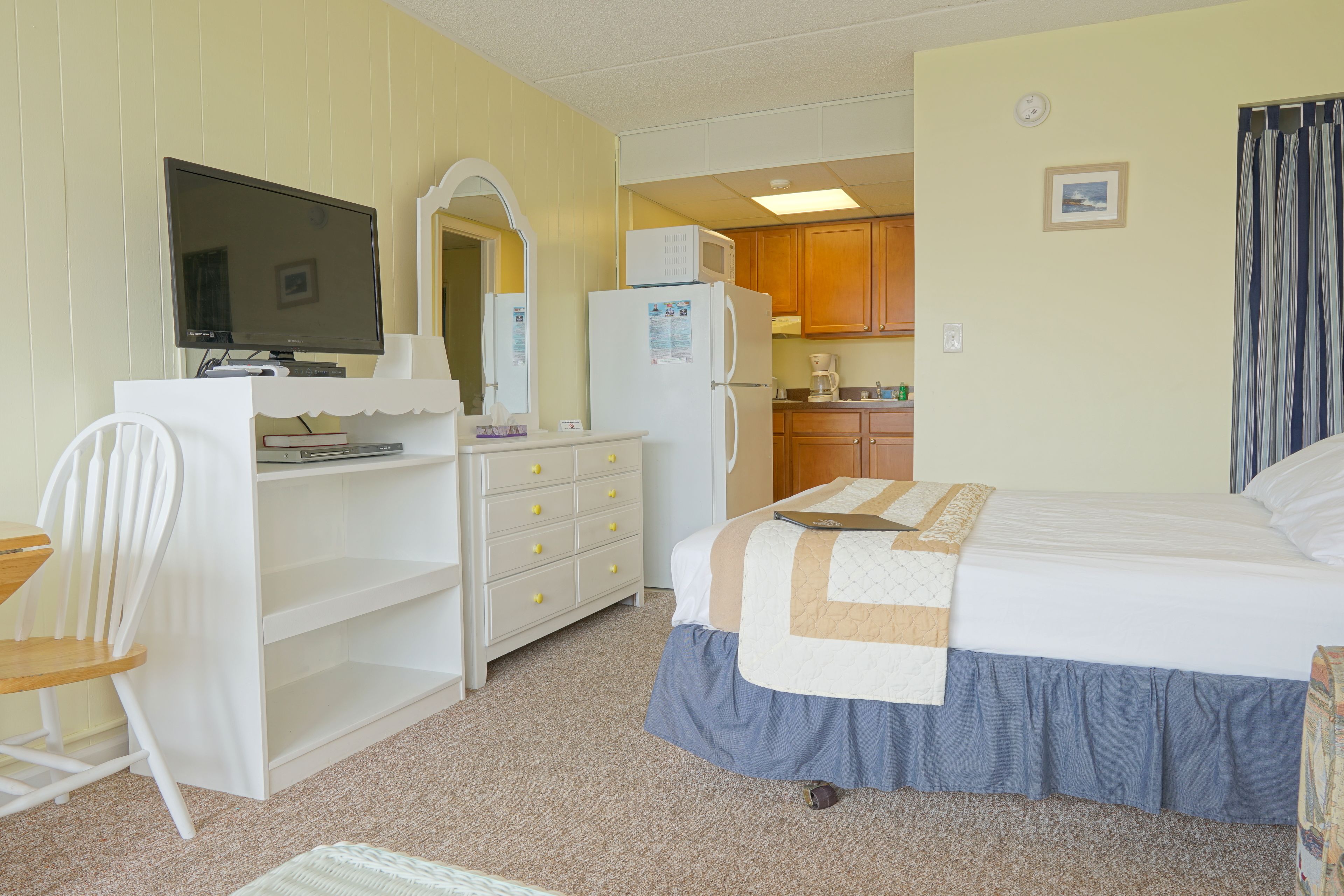 Une chambre avec un lit, un bureau avec un ordinateur, une chaise, une commode, un four à micro-ondes et un coin cuisine à l’arrière-plan.