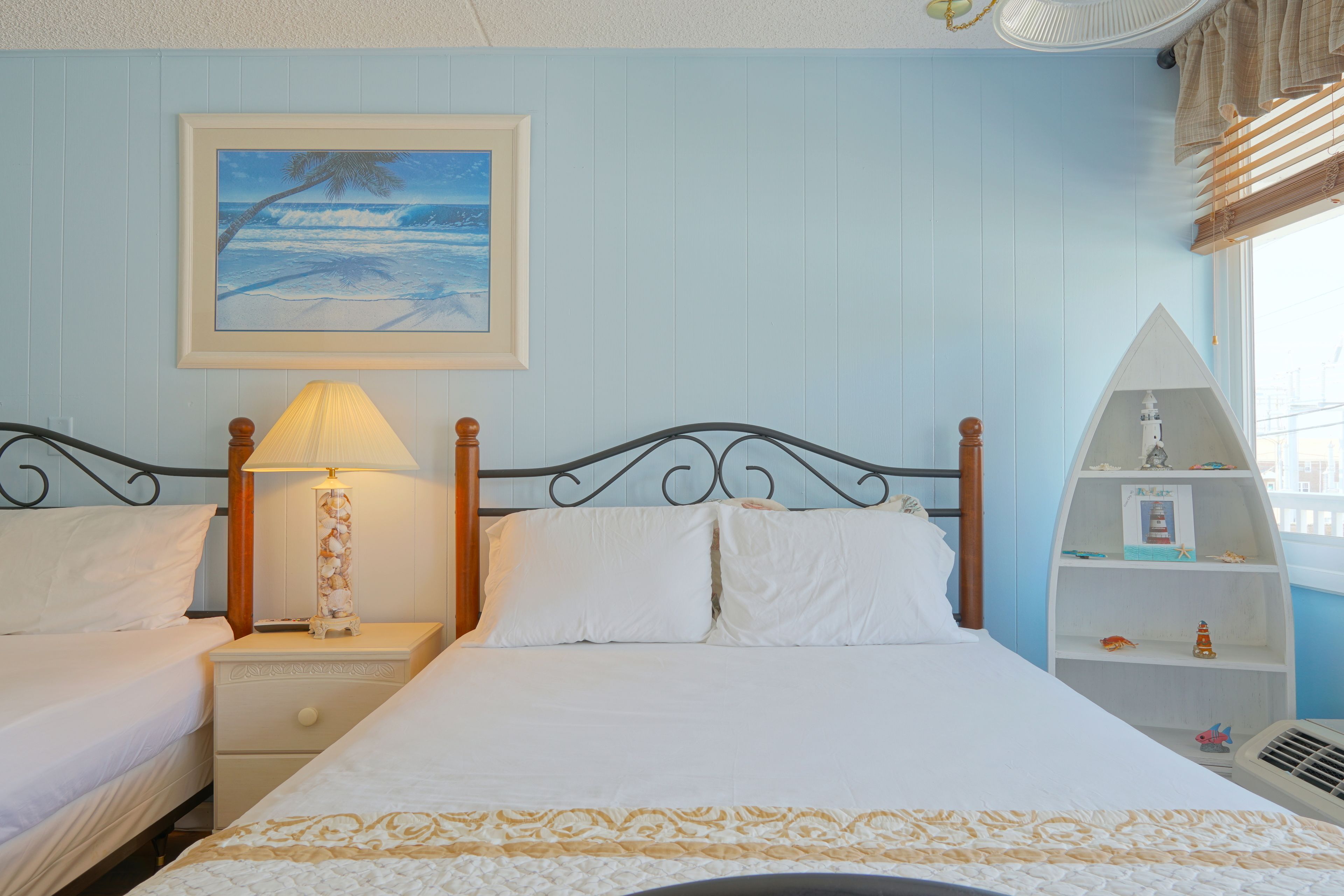 Une chambre avec deux lits, une table de chevet avec une lampe, une étagère avec des objets décoratifs et un tableau représentant une scène de plage.