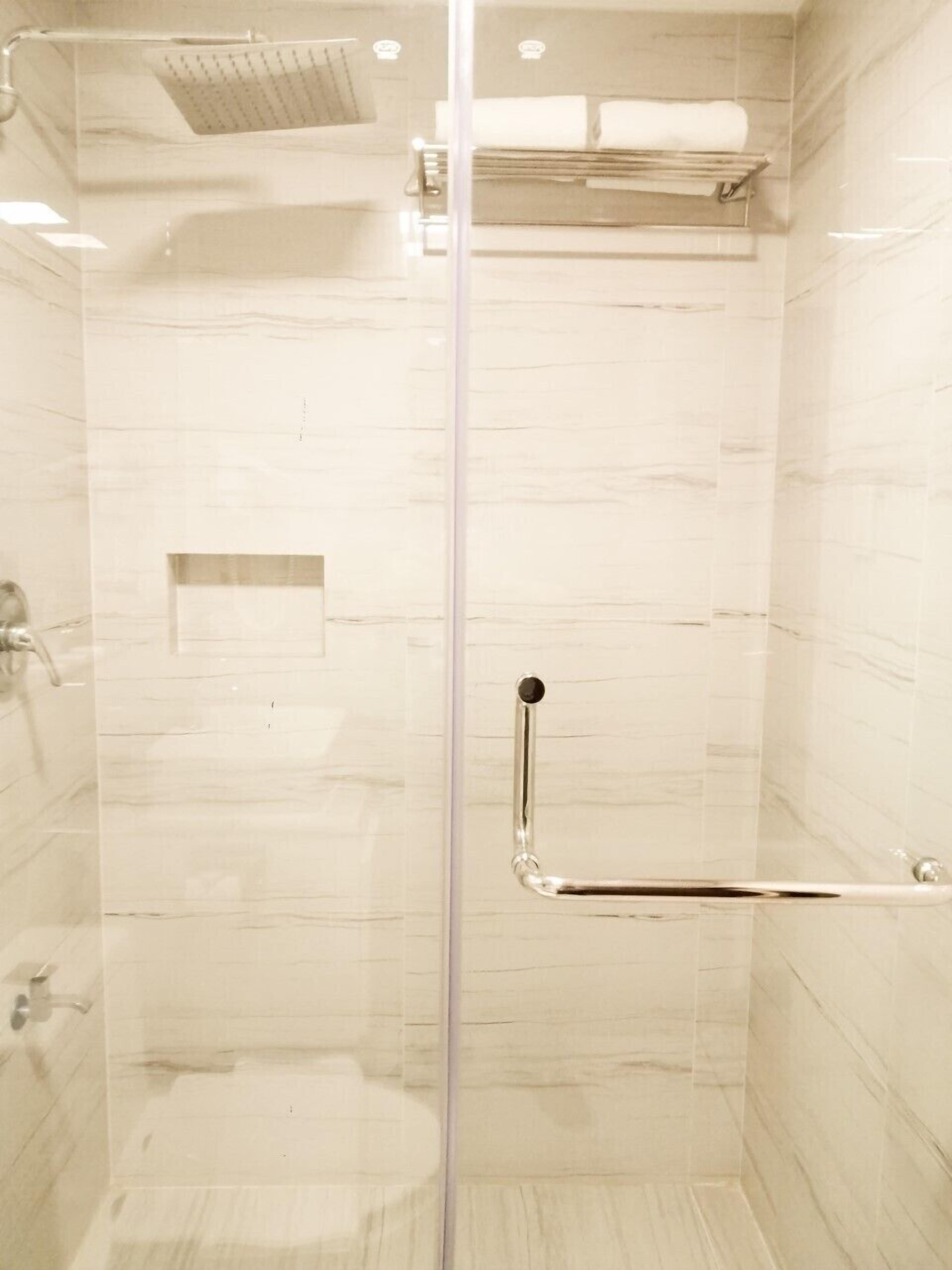 Cabina de ducha con paredes de vidrio, cabezal de ducha manual, toallero y un estante pequeño.