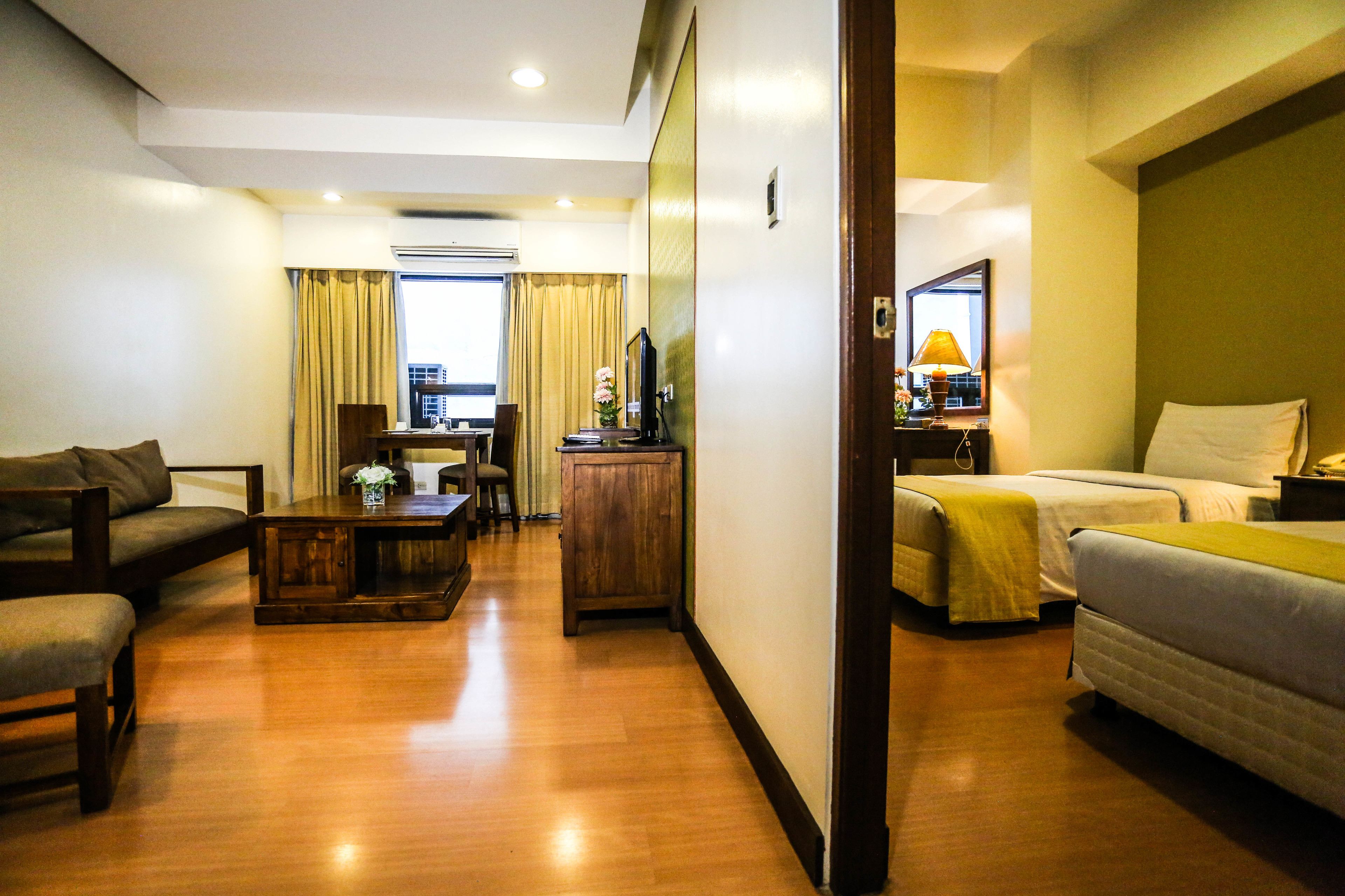 Habitación de hotel con piso de madera, un sofá, una cama, un escritorio con lámpara y vista a la ciudad a través de la ventana.
