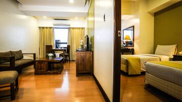Habitación de hotel con piso de madera, un sofá, una cama, un escritorio con lámpara y vista a la ciudad a través de la ventana.