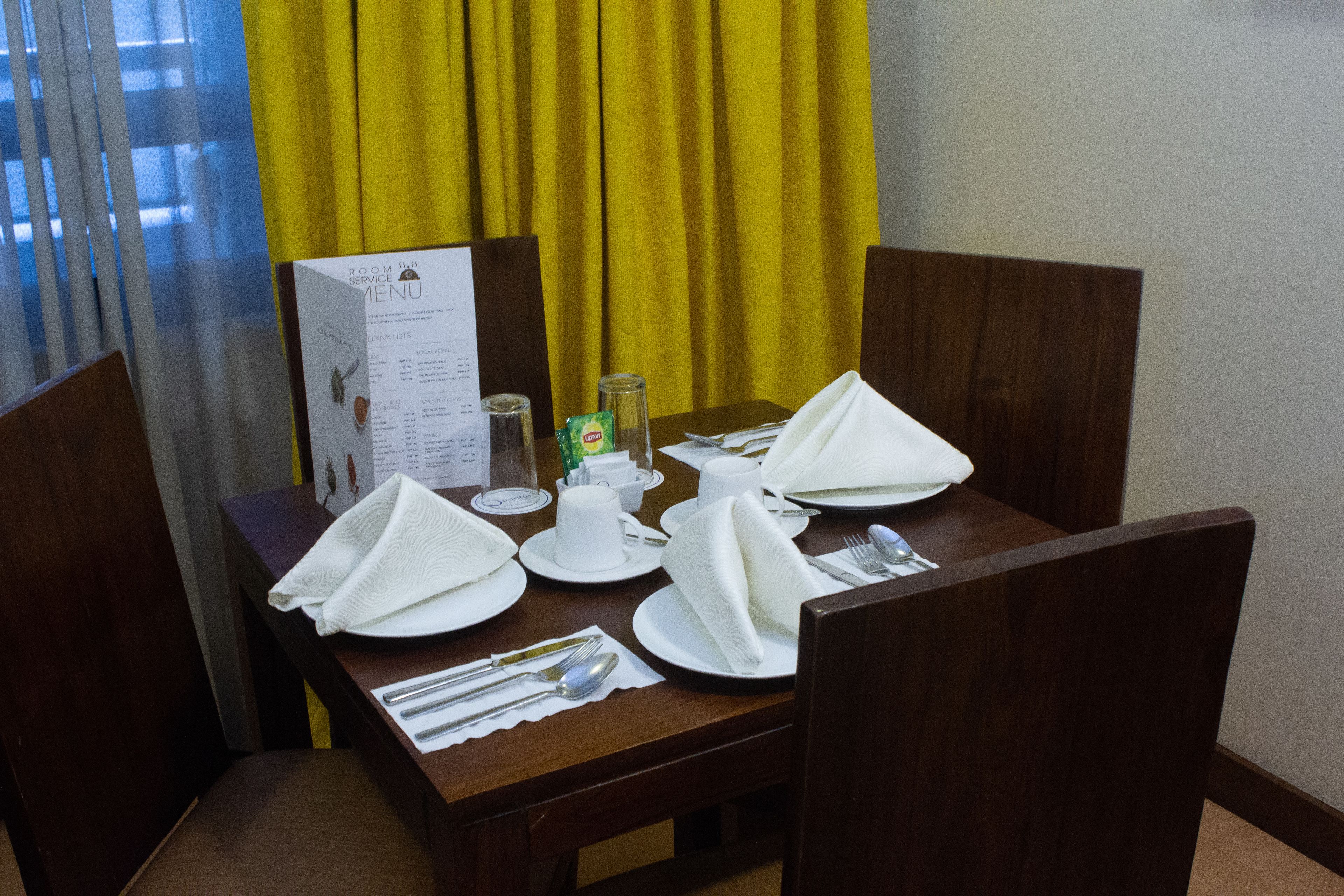 Une table de restaurant d’hôtel, dressée avec des serviettes blanches, des assiettes et des couverts, avec un présentoir à menus et des rideaux jaunes en arrière-plan.