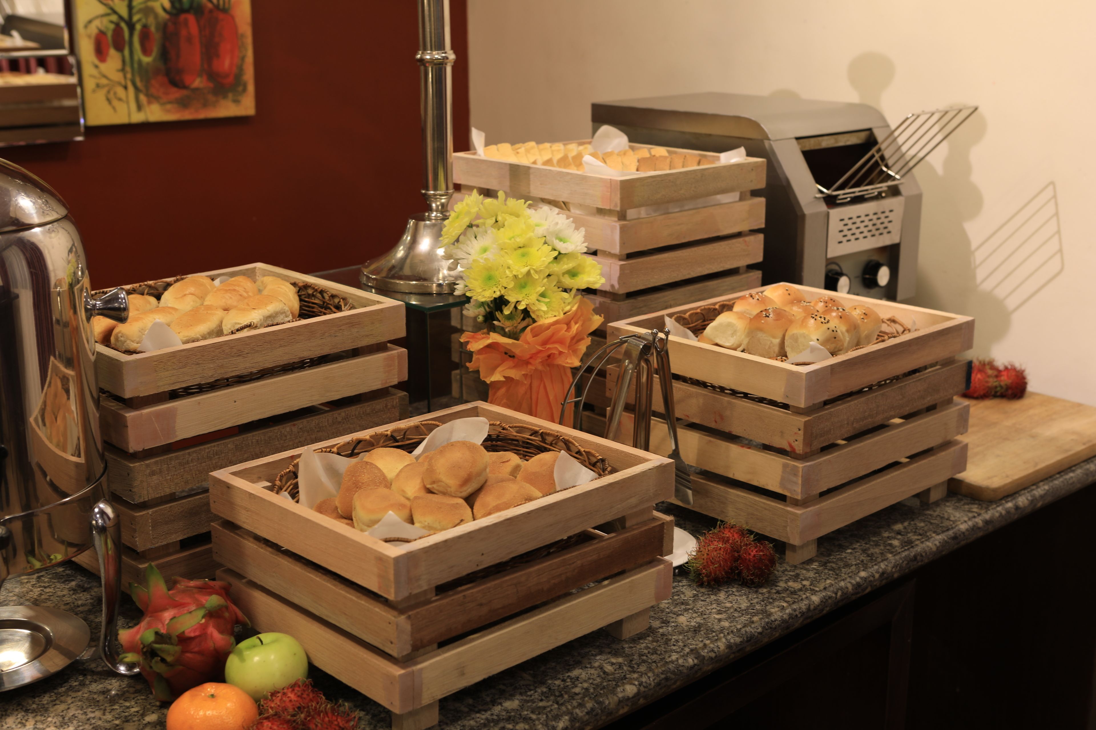 A buffet setup with various food items in wooden crates, a fruit bowl with dragon fruit and apples, and a decorative vase with flowers.