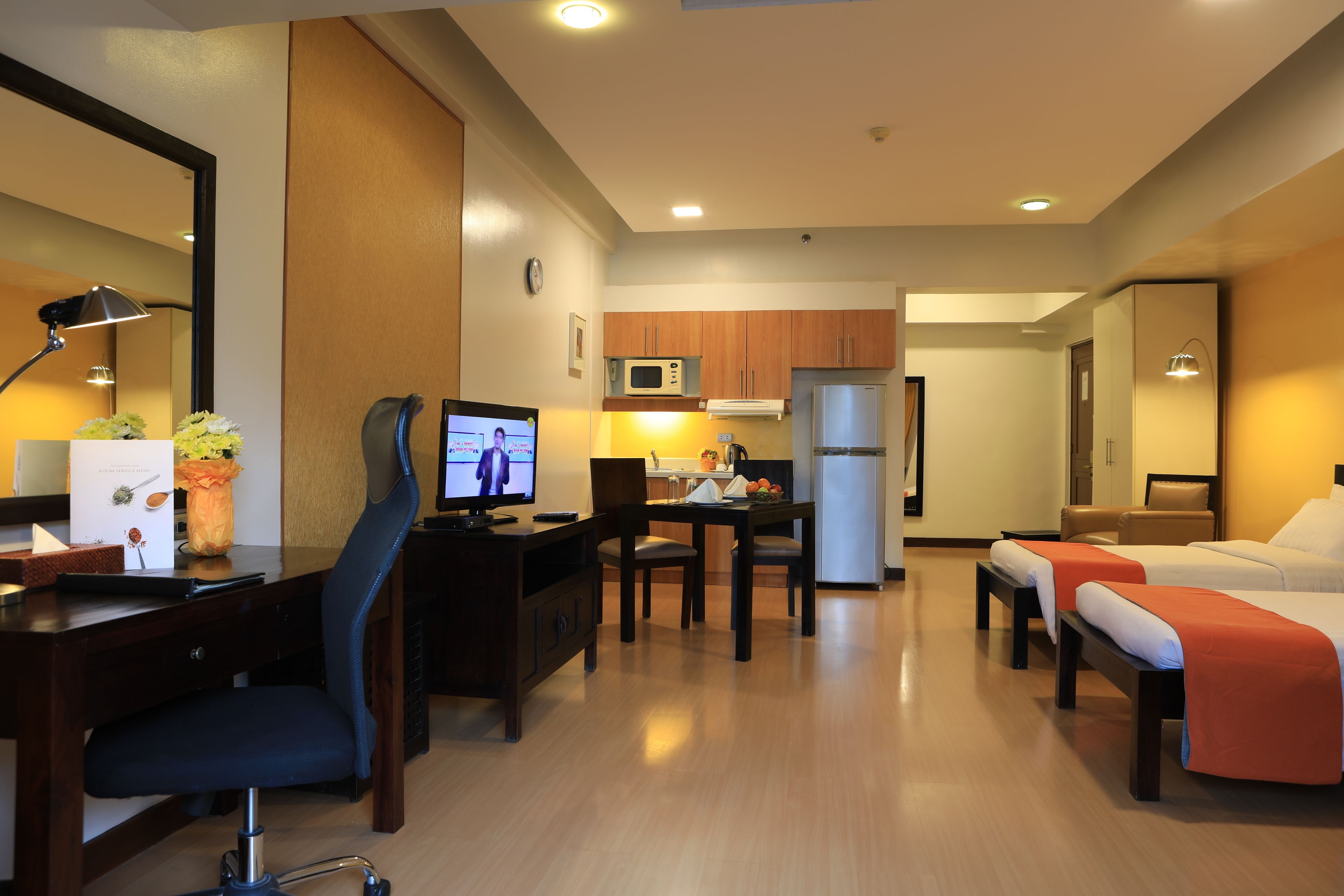 A hotel room with a desk, computer, chair, two beds, a small kitchenette, and a mirror.