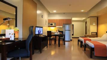 Habitación de hotel con escritorio, computadora, silla, dos camas, una kitchenette pequeña y un espejo.