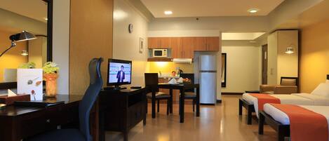 A hotel room with a desk, computer, chair, two beds, a small kitchenette, and a mirror.