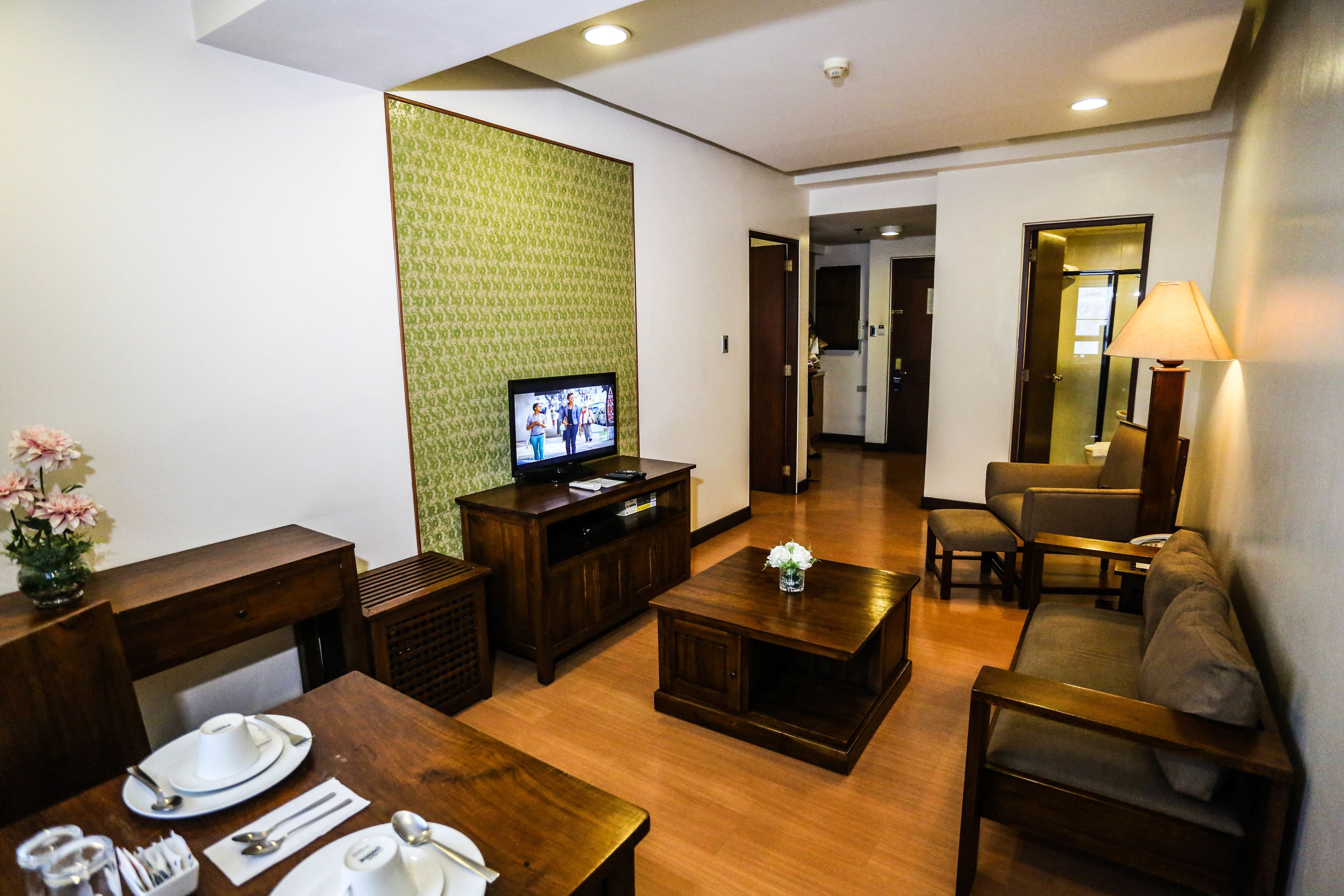 A hotel room with a wooden dining table set, a sofa, a television, and a green patterned wall.