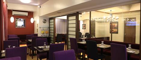 Un restaurante con sillas y mesas moradas, y un candelabro. Hay separadores de vidrio y una pared roja con un reloj.