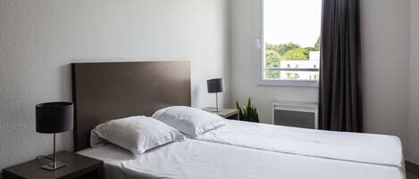 A hotel room with a bed, a nightstand, a lamp, a potted plant, and a window with curtains.