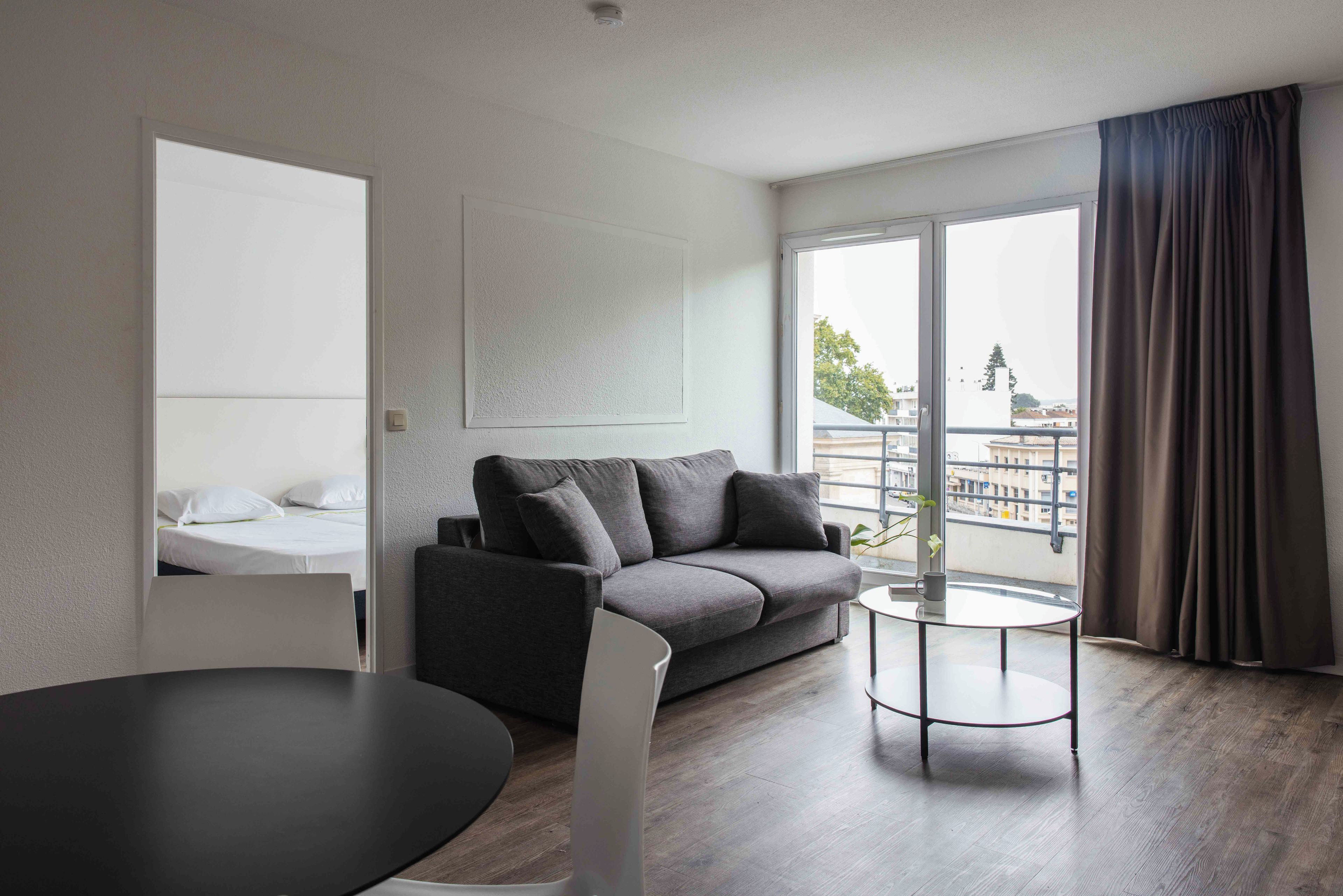 Un salon moderne avec un canapé gris, une table ronde et une grande fenêtre avec des rideaux.