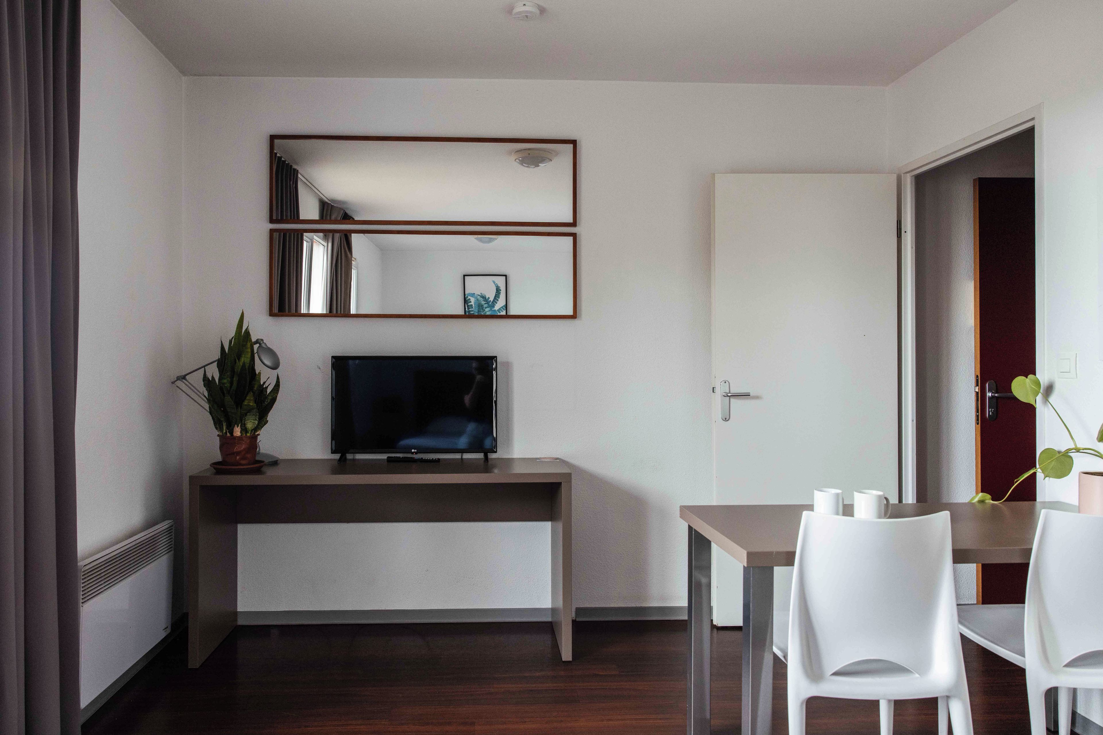 A hotel room with a desk, a television, a potted plant, and a dining area with chairs.