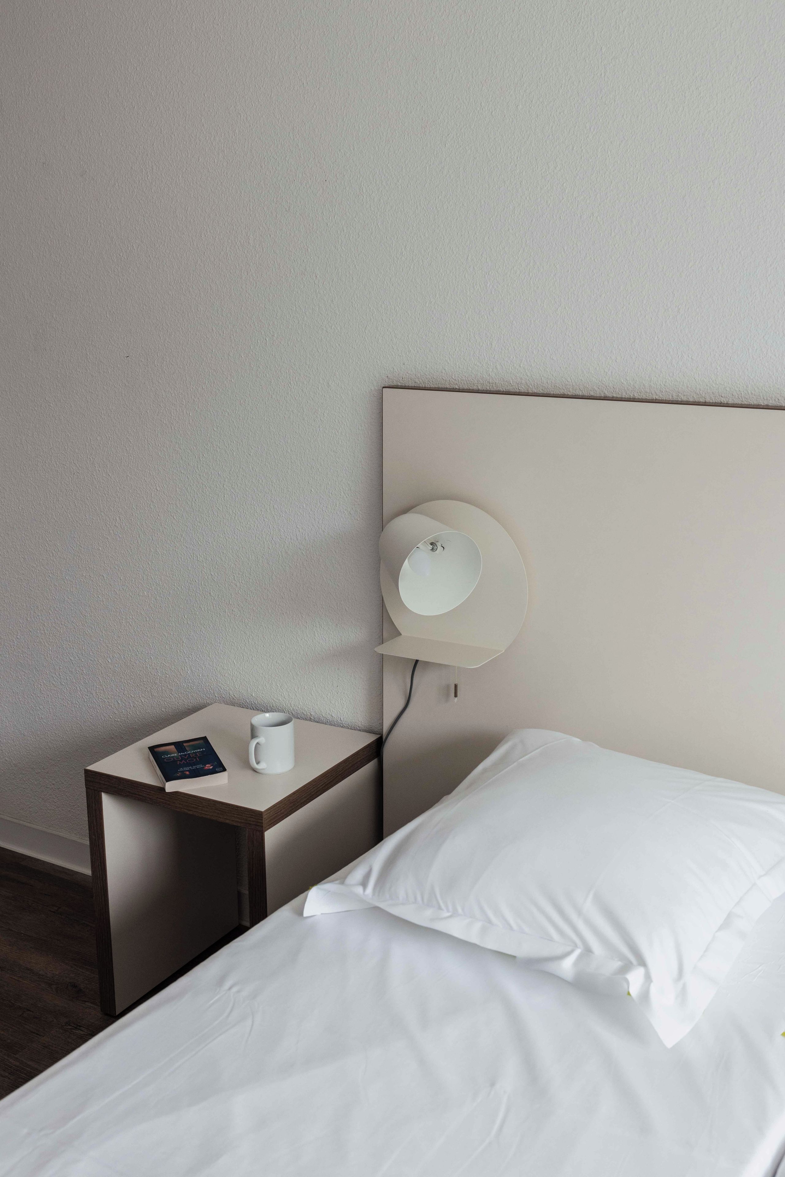 A hotel room with a single bed, a bedside table with a lamp, a cup, and a book.