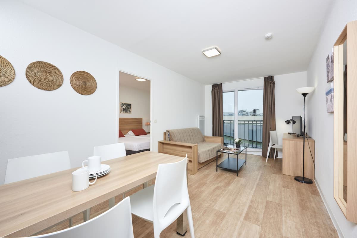 Quarto de hotel moderno com uma mesa de jantar de madeira, cadeiras brancas, um sofá, uma pequena mesa de centro e uma varanda com vista.