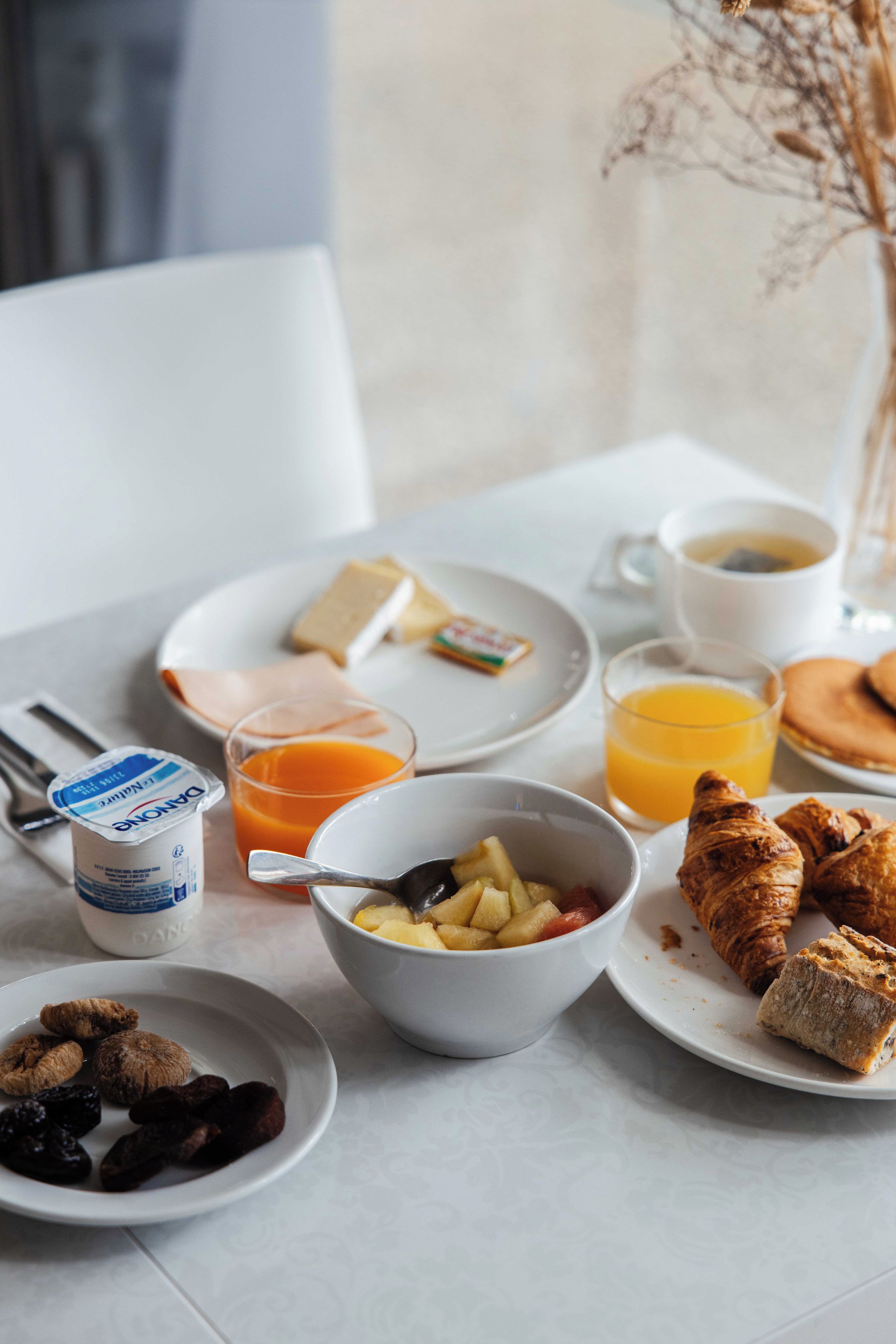 Une table mise pour le petit-déjeuner avec une assiette de croissants, un bol de fruits, un verre de jus d’orange, une tasse de café et un pot de yaourt.