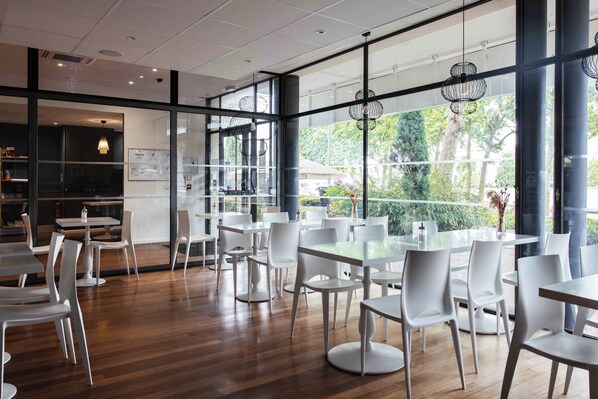 Interior de un restaurante moderno con mesas y sillas blancas, ventanas amplias y vistas a zonas verdes.