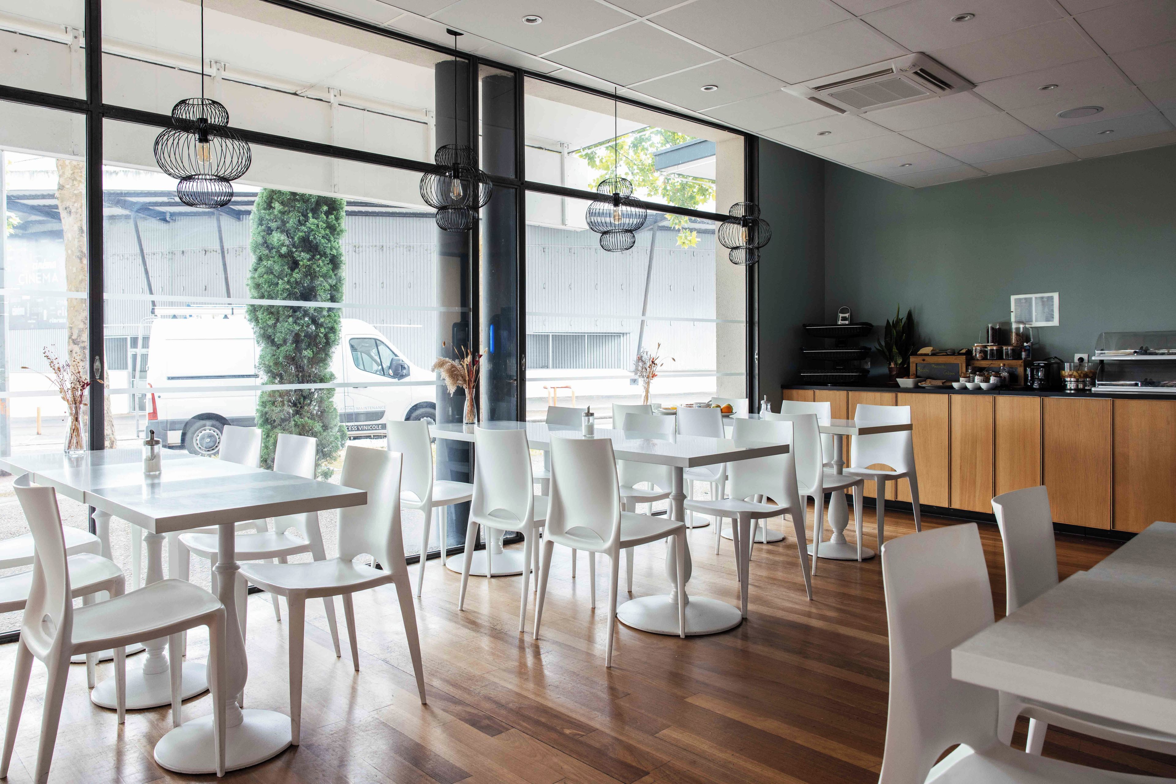 A modern cafe interior with white tables and chairs, large windows, and a wooden counter.