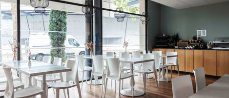 A modern cafe interior with white tables and chairs, large windows, and a wooden counter.