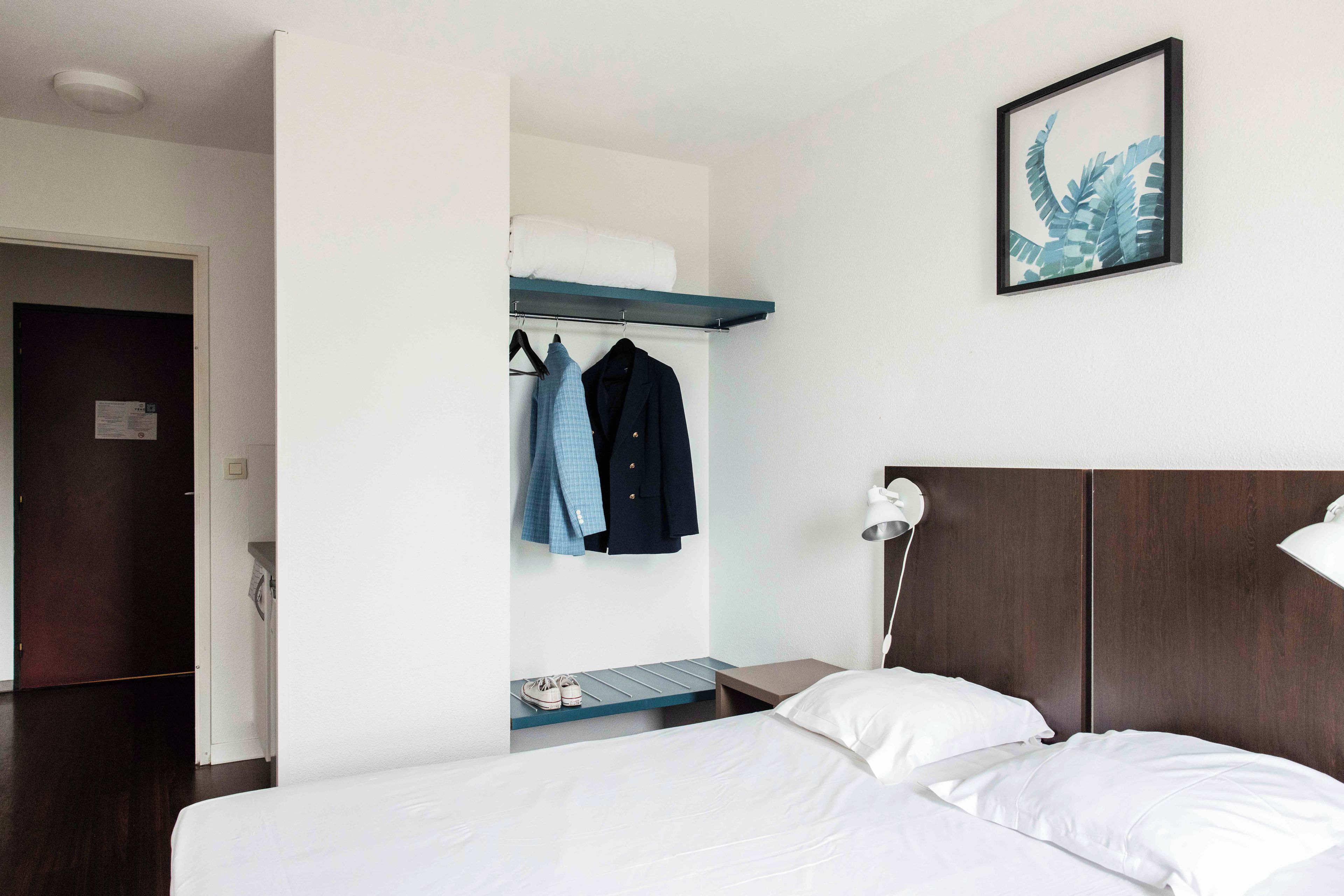 A hotel room with a bed, a nightstand, a wall-mounted shelf with clothes, a framed picture, and a door leading to another room.