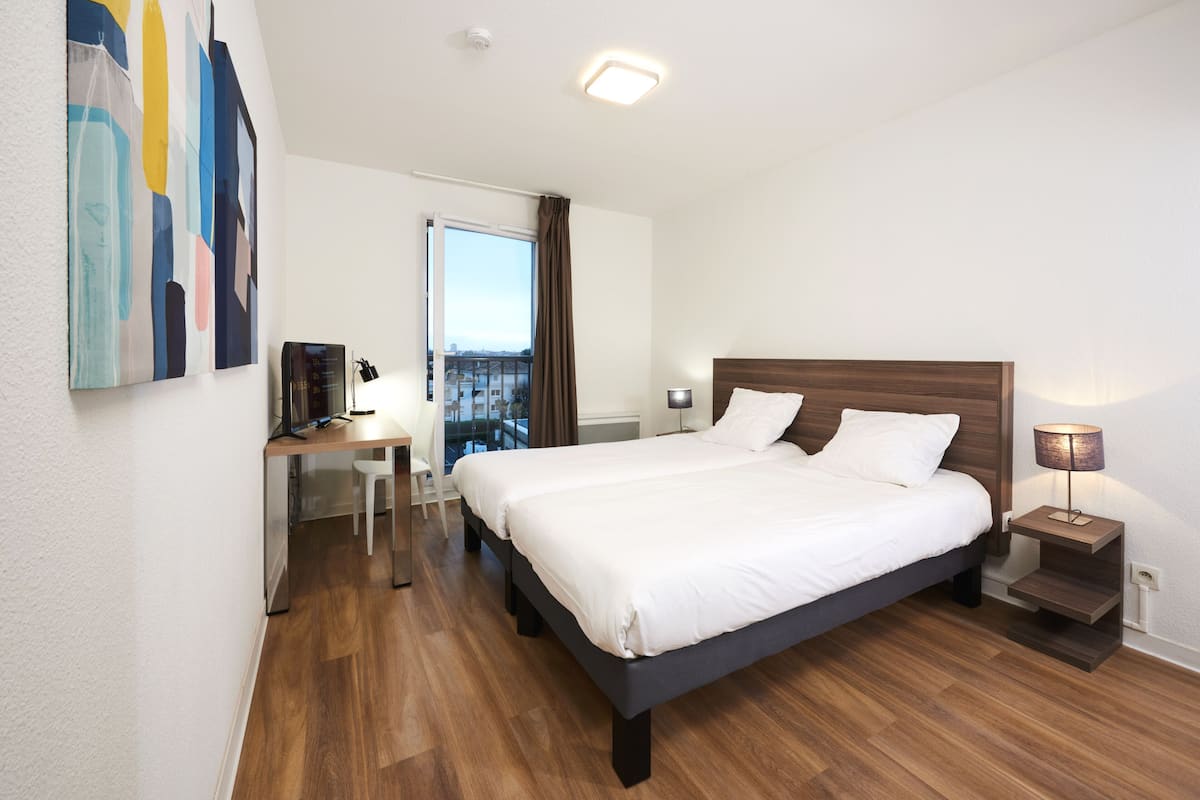 Quarto de hotel moderno com uma cama grande, uma escrivaninha com cadeira, uma mesinha e vista para a cidade pela janela.