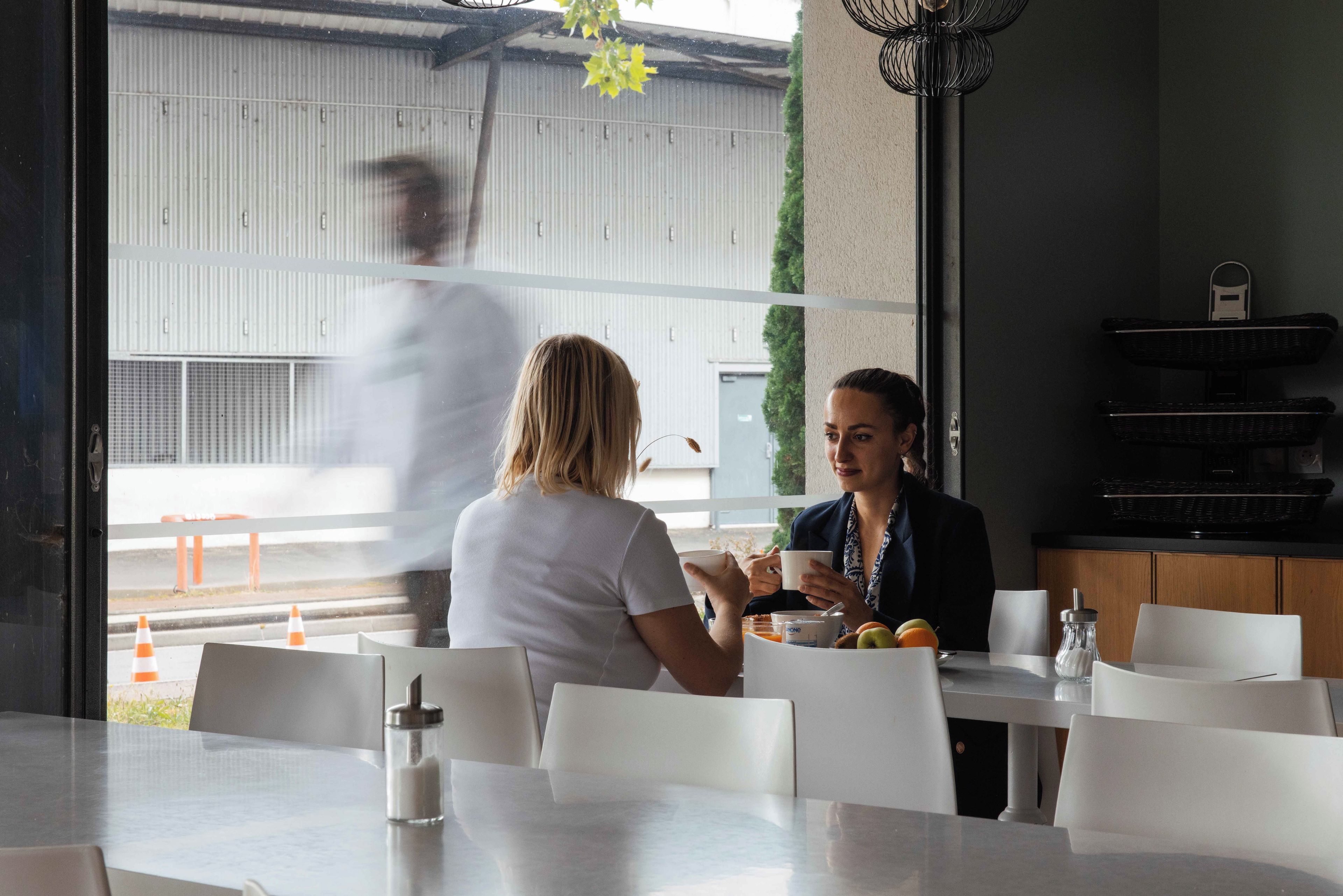 Un comedor con una mesa dispuesta para dos, vista al exterior y una persona caminando afuera.