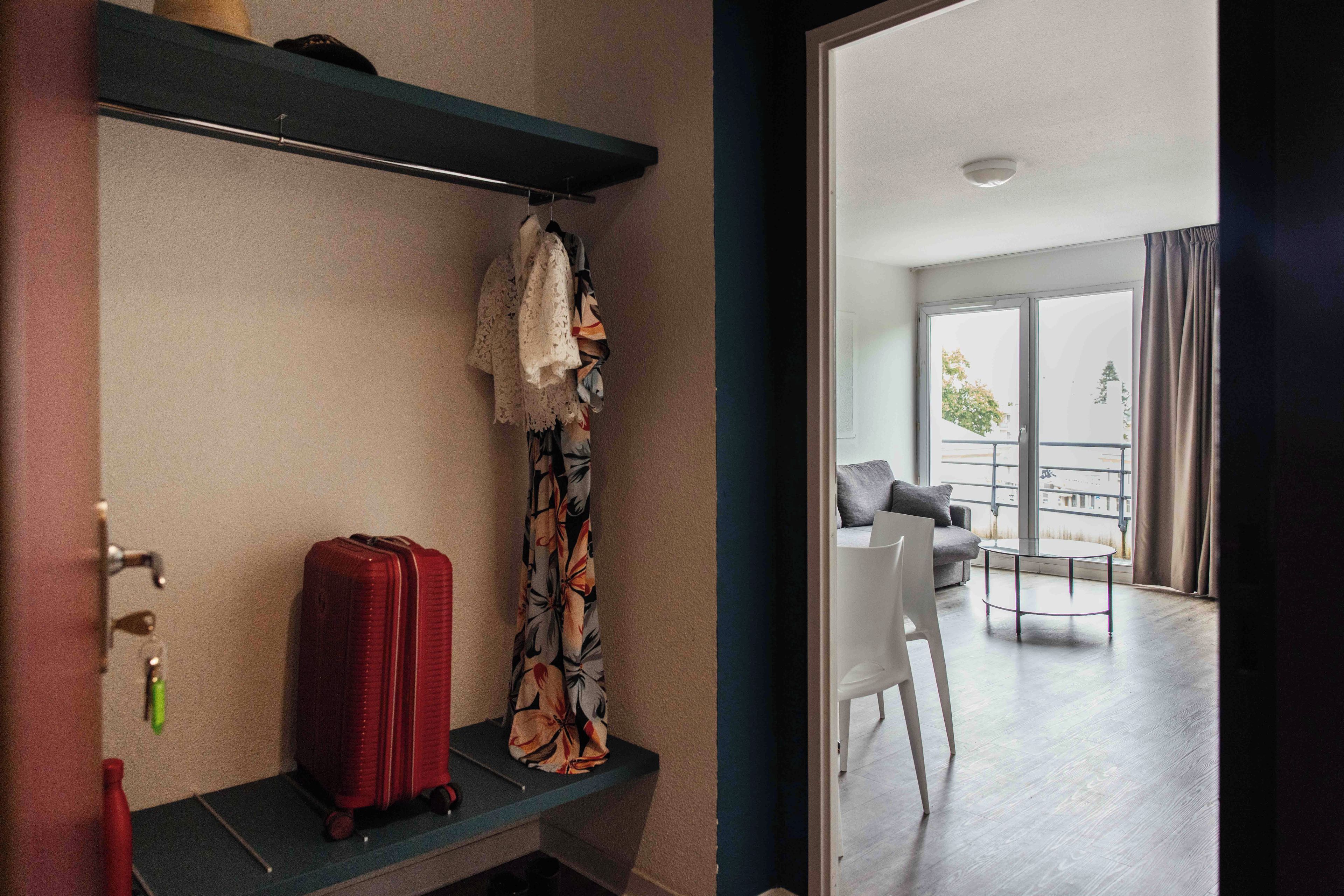 Una habitación de hotel con un banco, una maleta roja y una cortina estampada.