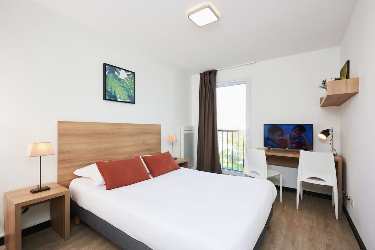 Quarto de hotel com cama, escrivaninha, televisão e janela com cortinas.