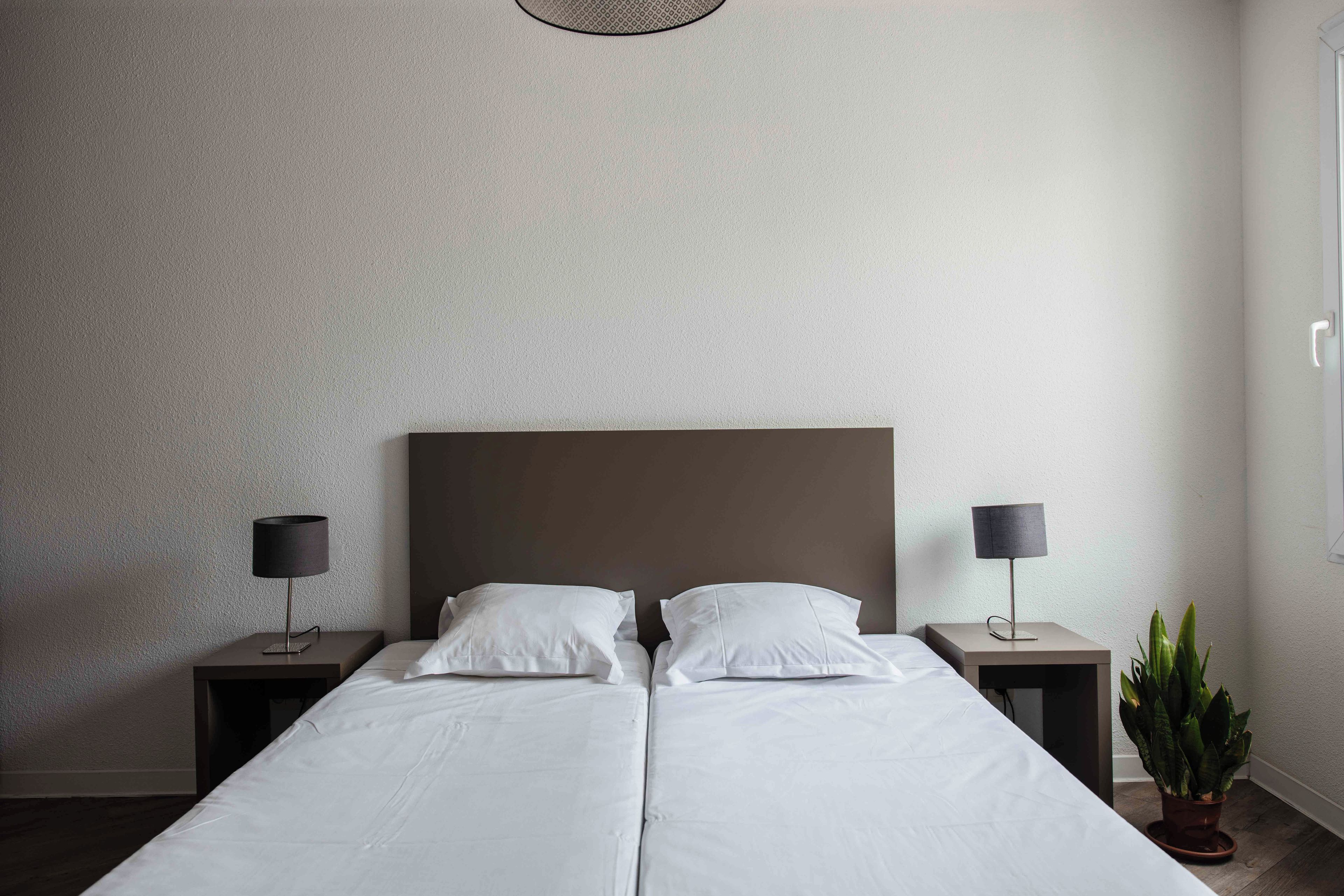 A double bed with white linens, two bedside tables with lamps, and a potted plant on the right side.
