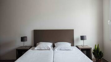 A double bed with white linens, two bedside tables with lamps, and a potted plant on the right side.