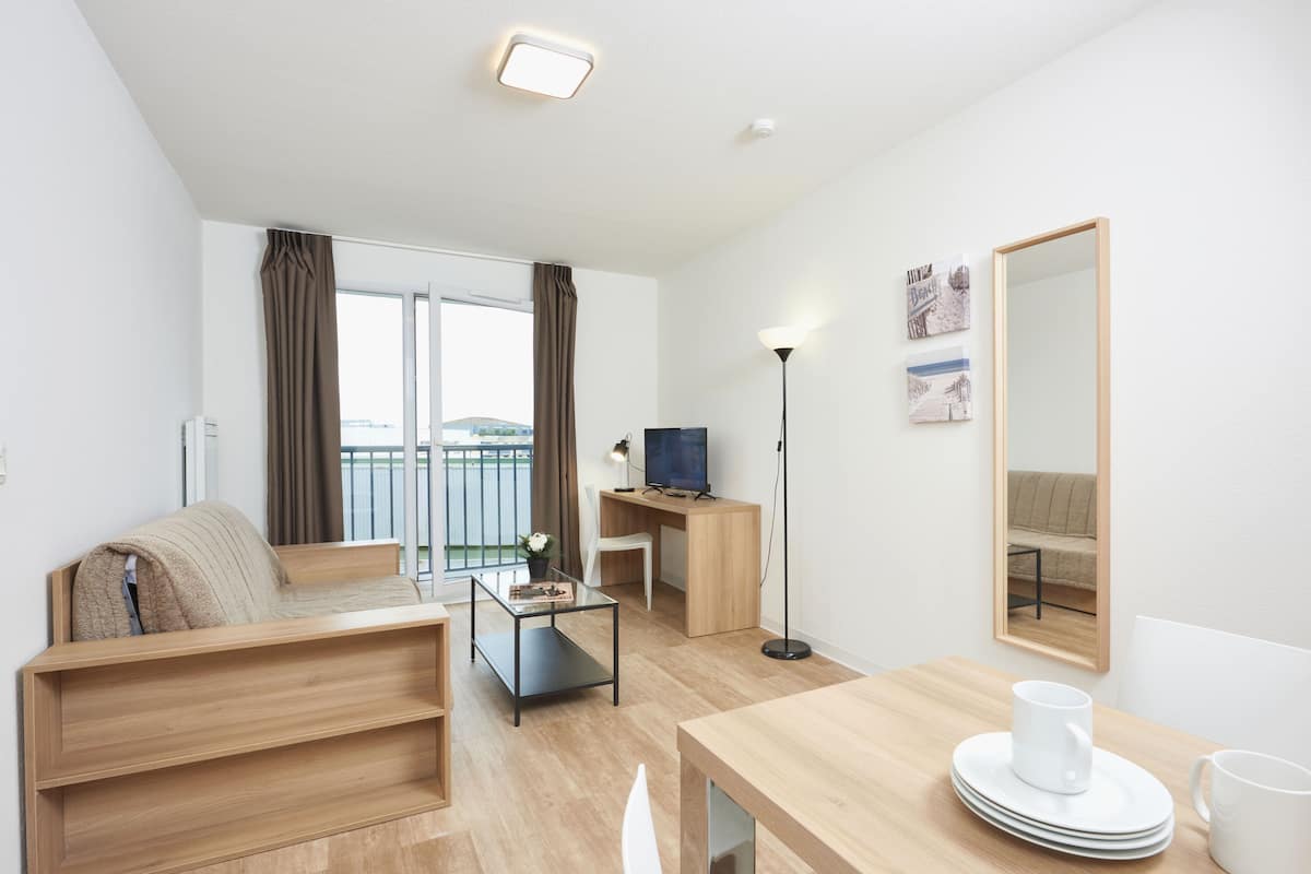 Quarto de hotel moderno com sofá, escrivaninha, mesa de jantar e uma grande janela com cortinas.