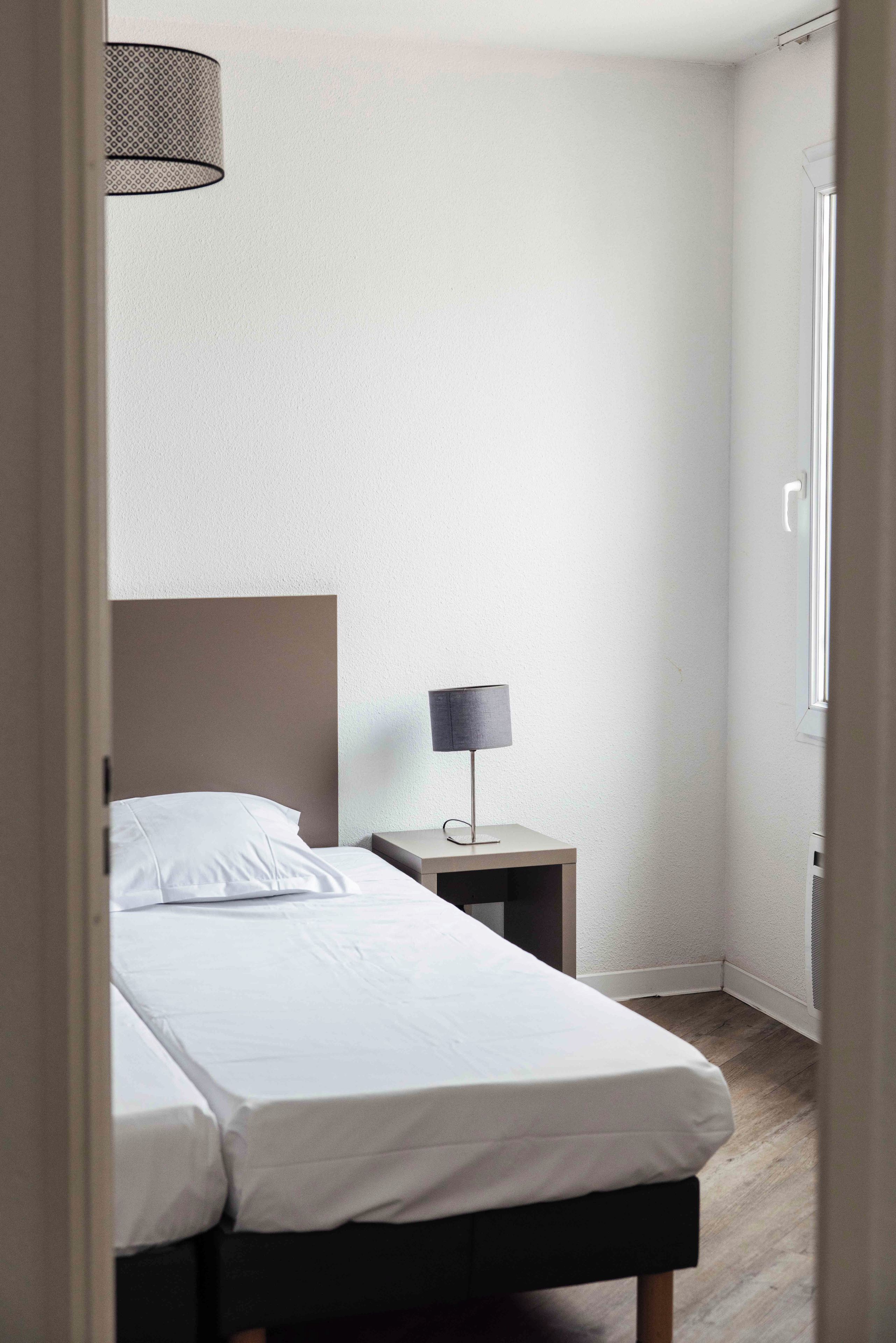 A small, neatly made bed with white linens in a room with a plain wall and a window.