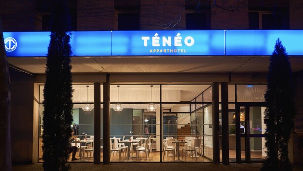 The exterior of the Teneo Apparthotel with a modern design, featuring glass doors and a blue illuminated sign. - Ténéo Apparthotel Talence - Centre (Talence)