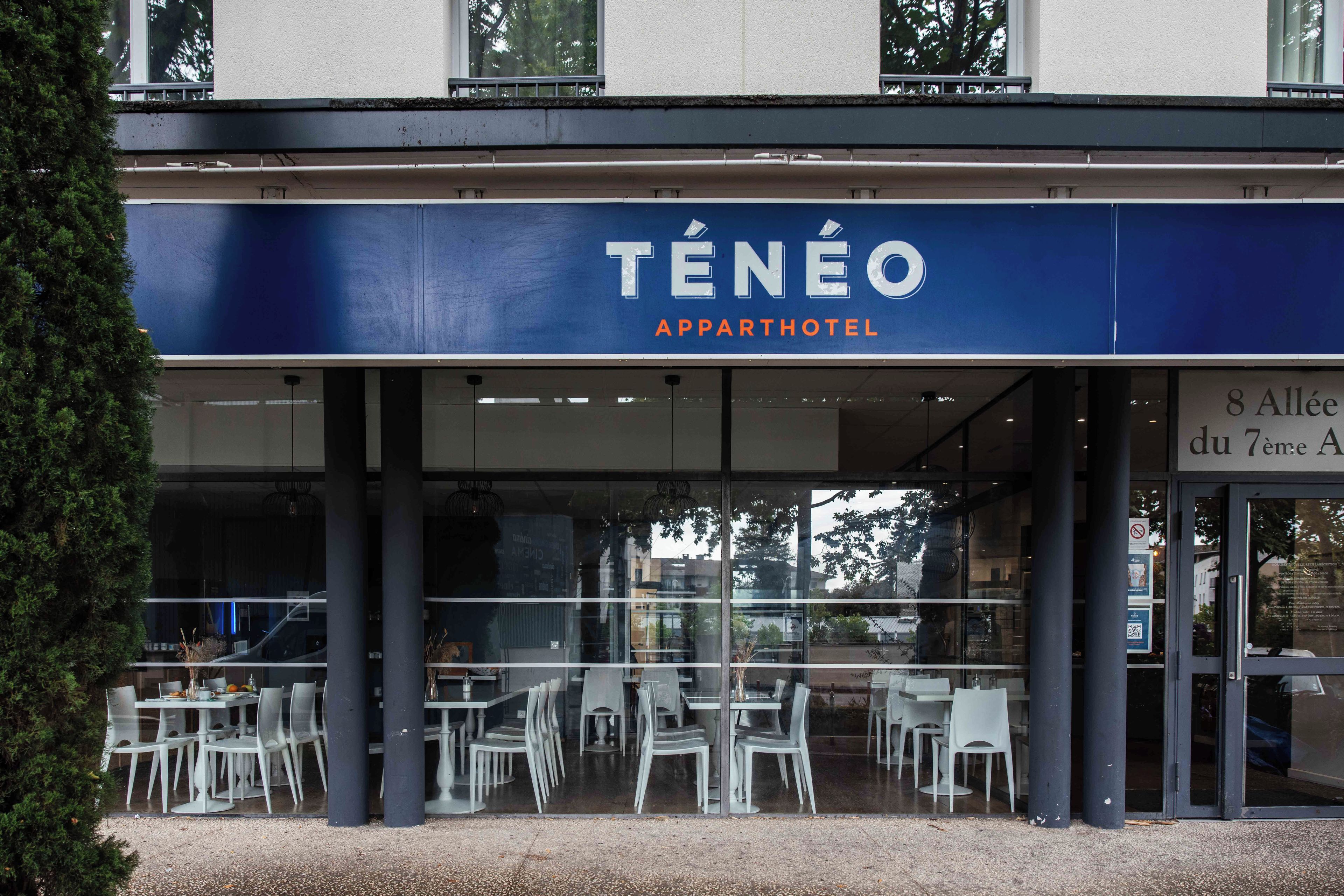 An outdoor seating area of a Ténéo Apparthotel with white chairs and tables, and a view of the street.