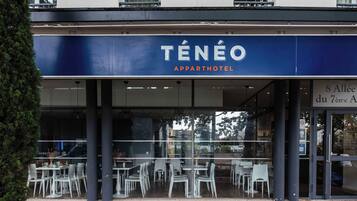 An outdoor seating area of a Ténéo Apparthotel with white chairs and tables, and a view of the street.
