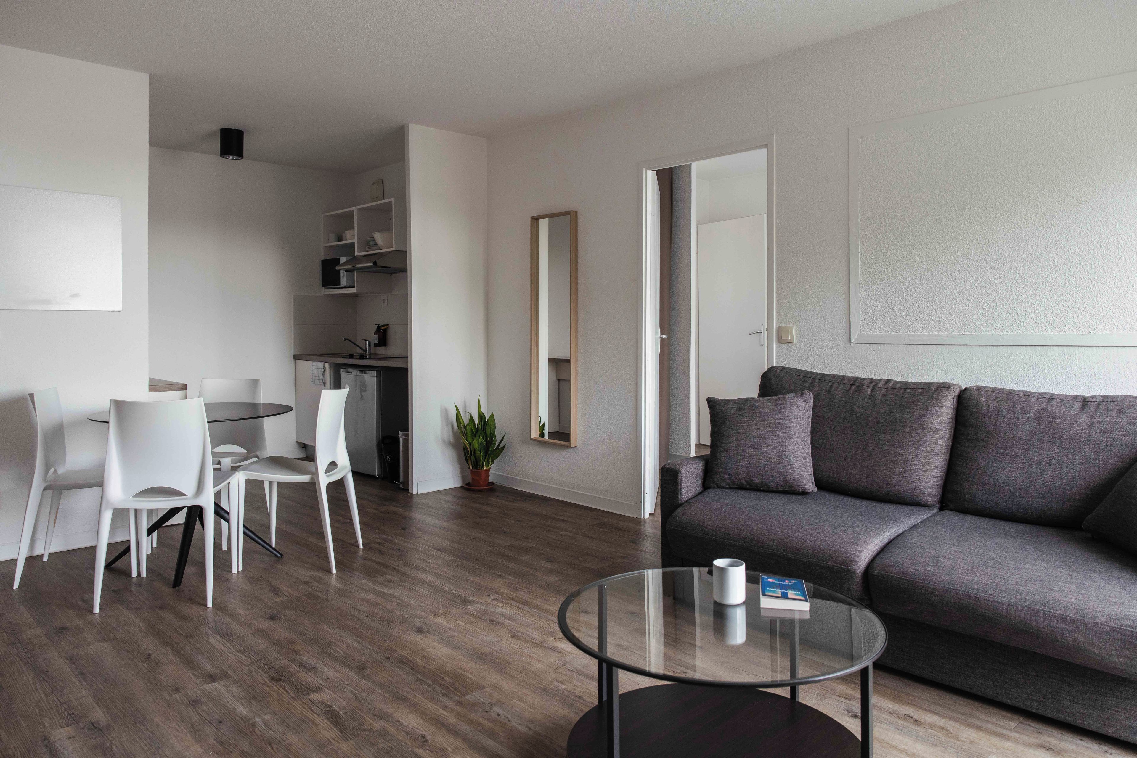 Ein modernes Wohnzimmer mit einer grauen Couch, einem Glastisch und einem Essbereich mit weißen Stühlen.