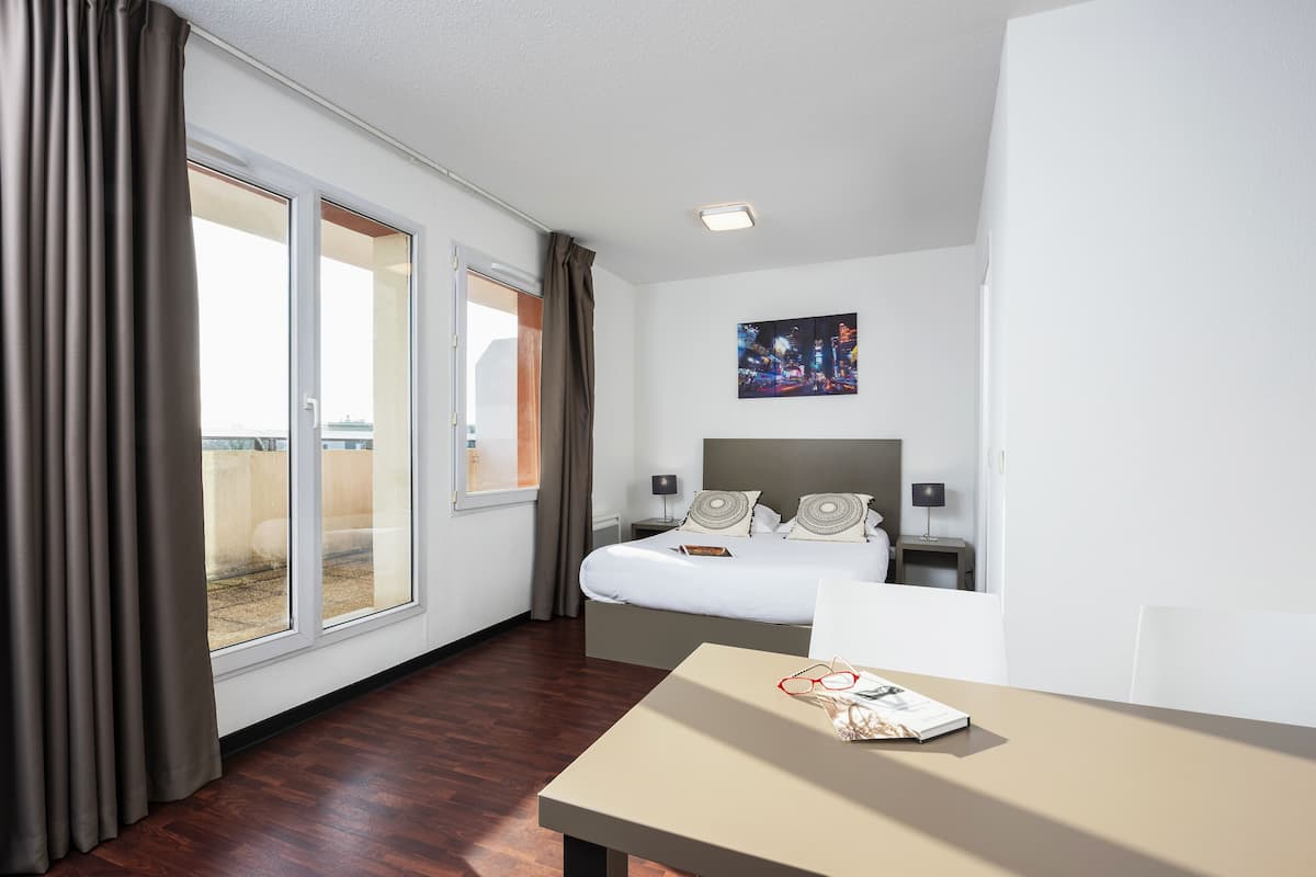 Quarto de hotel com cama, mesa, mesa de cabeceira, um quadro e vista através de uma porta de correr.