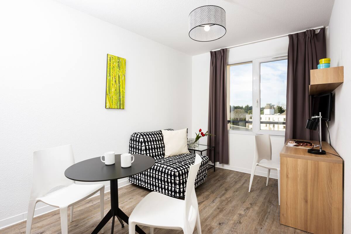 Quarto moderno com sofá estampado em preto e branco, uma pequena mesa redonda com duas cadeiras brancas, mesa lateral de madeira com abajur e uma grande janela com cortinas.
